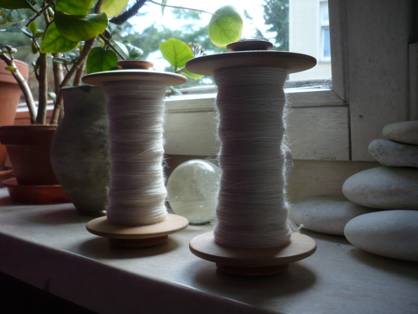 two bobbins with natural white handspun before the window, surrounded by some plants, minerals and stones