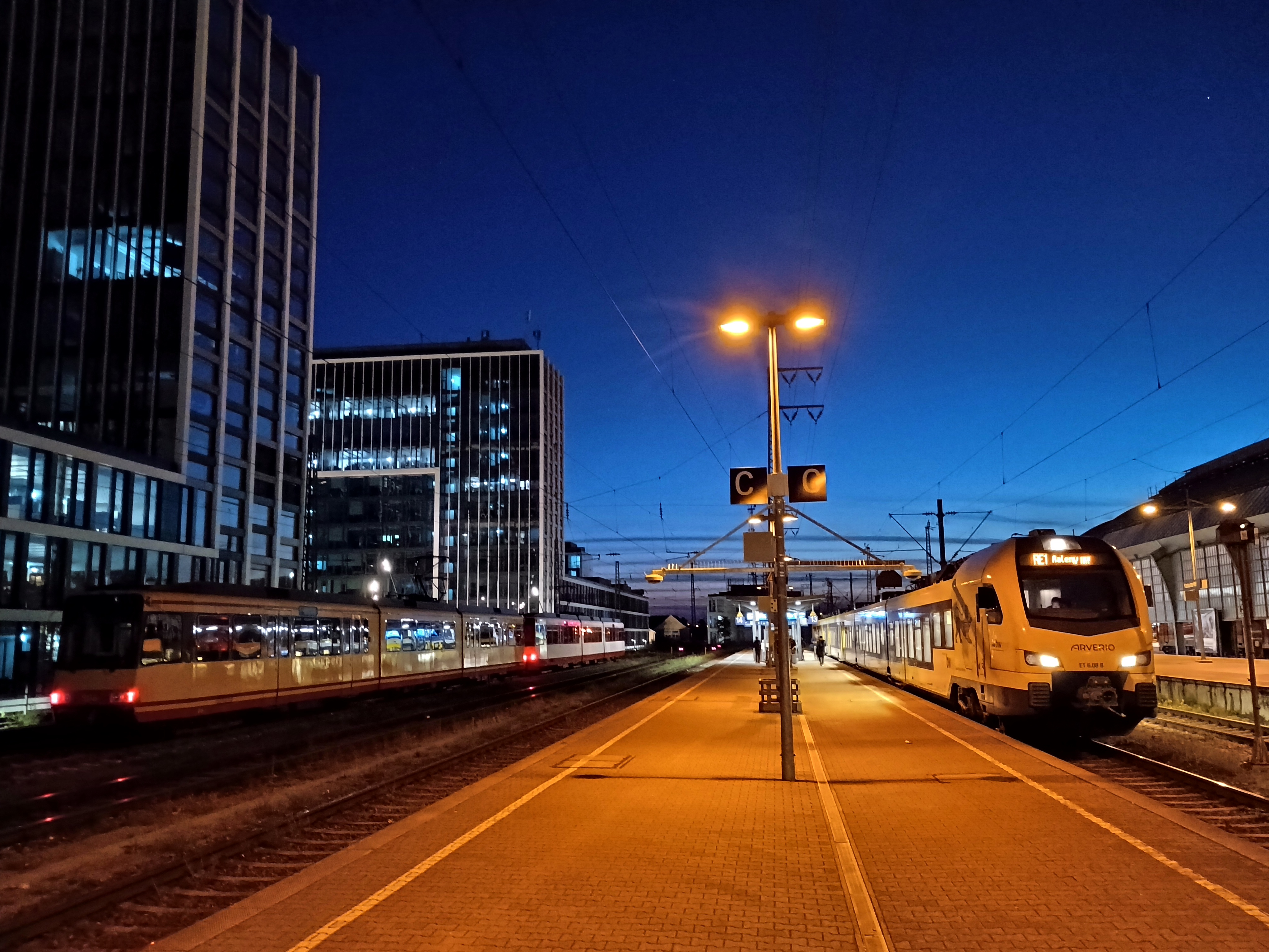 Abendstimmung auf Gleis 13/14 am Hauptbahnhof Karlsruhe. Der Horizont hat einen blauen Farbverlauf durch die Abenddämmerung, der Bahnsteig ist orange beleuchtet. Links stehen vor den Bürogebäuden zwei abgestellte Stadtbahnwagen. Rechts wartet ein Regionalexpress auf Abfahrt.