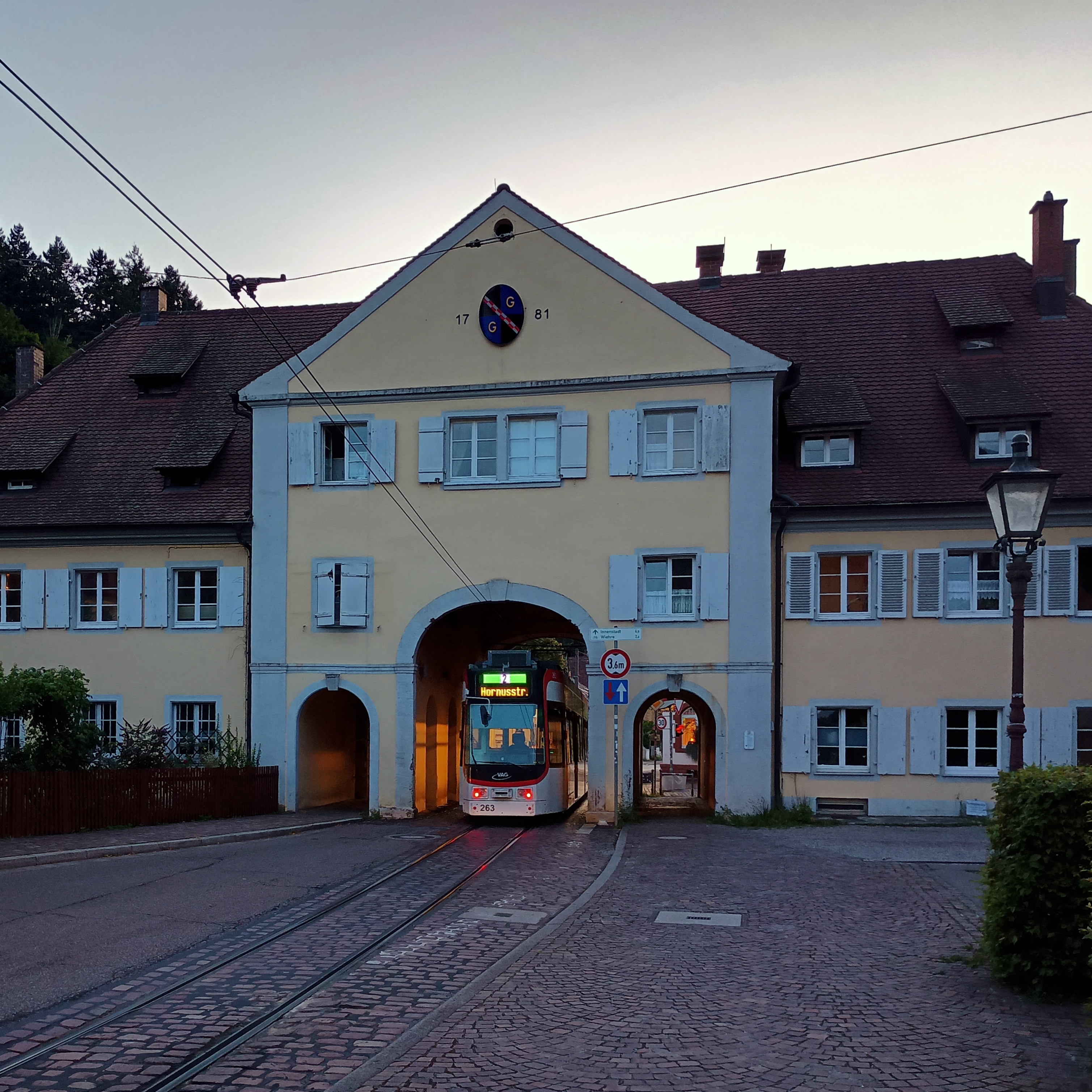 Eine Tram der Linie 2 zur Hornusstraße fährt im letzten Licht der Abendsonne durch das Tor in Freiburg-Günterstal, von hinten aufgenommen.