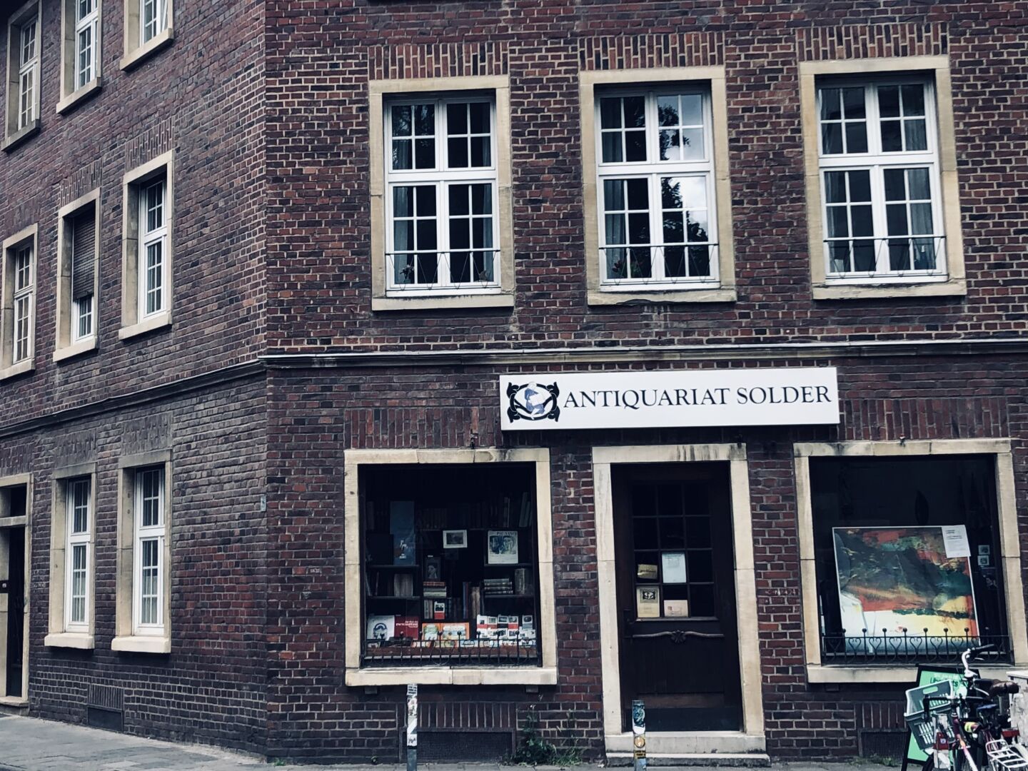 Ein Backstein-Eckhaus mit einem Antiquariat in der unteren Etage. Zwei kleine Schaufenster mit Tür dazwischen. Darüber eine Reihe schmale unterteilte Fenster. (Das Geschäft ist Schauplatz in „Wilsberg“-Krimireihe.)