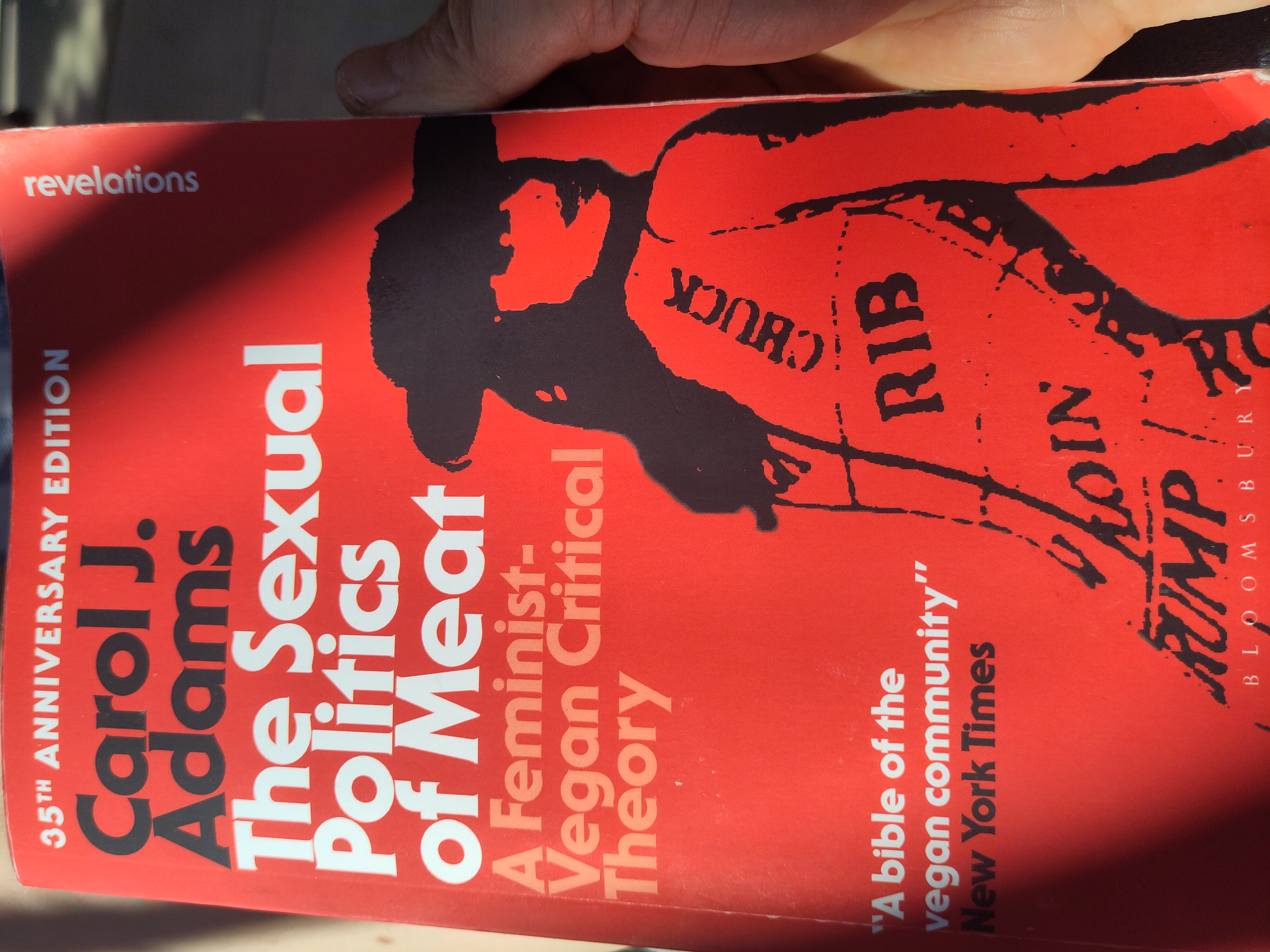 Close-up photo of the bright red cover of the book *The Sexual Politics of Meat* by Carol J. Adams, shown in warm sunlight and partly held by a hand on the right side. The cover features bold black and white typography and a black silhouette illustration of a woman whose body is marked with butcher-style meat cuts labeled “RIB,” “LOIN,” and “CHUMP.” The subtitle reads *A Feminist-Vegan Critical Theory*. A quote from the *New York Times* calls it “A bible of the vegan community.”