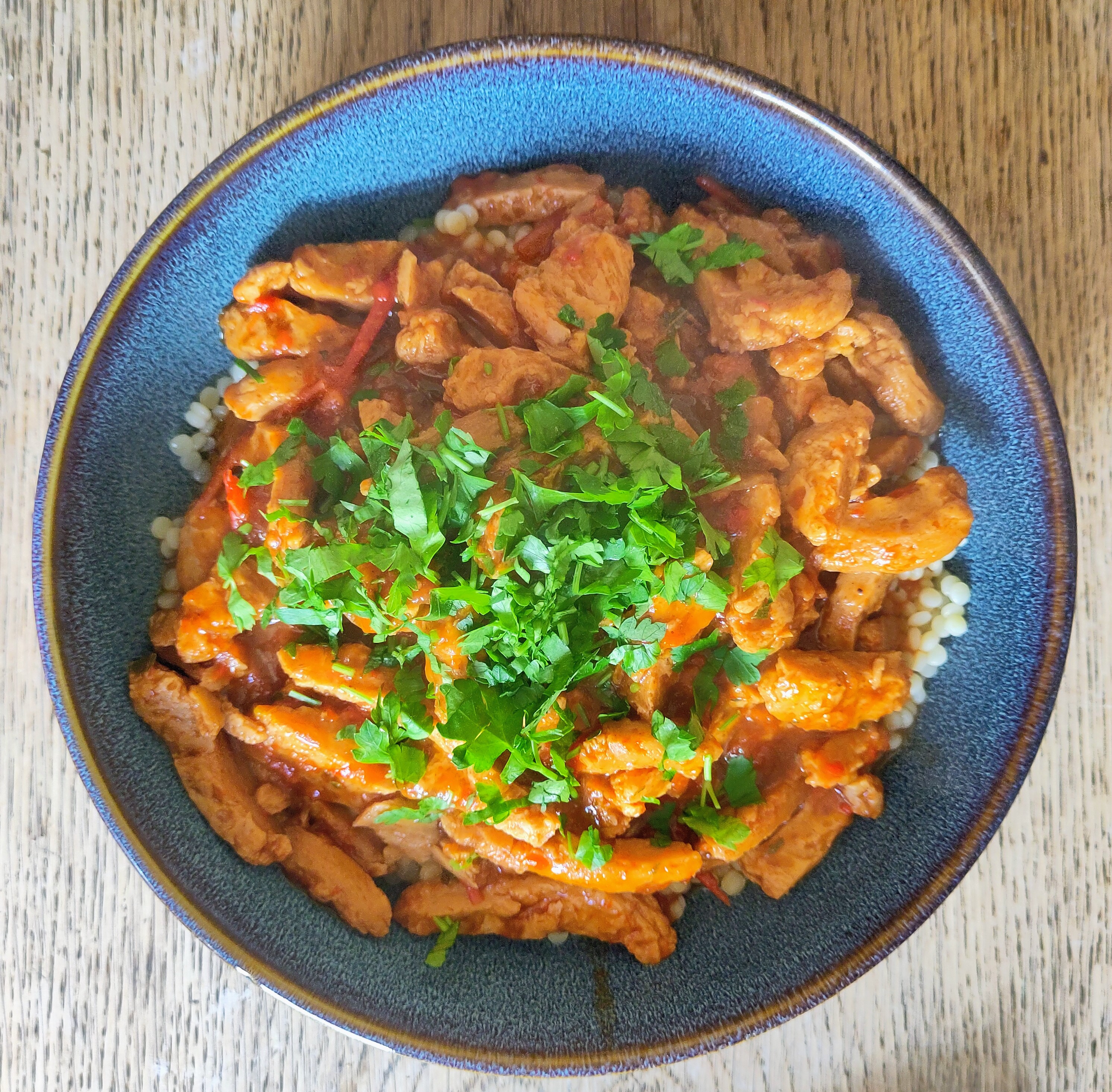 Ein dunkelblauer Teller steht auf einem Holztisch. Darauf liegt ein Gericht aus kleinen Nudeln, bedeckt mit Sojastreifen in einer rötlich-orangefarbenen Sauce. Zwischen dem Sojastreifen sind Stücke von Paprika und Zwiebeln zu erkennen. In der Mitte liegt eine großzügige Portion frisch gehackter grüner Petersilie als Topping. Das Essen wirkt warm und frisch gekocht, leichter Dampf steigt auf.
