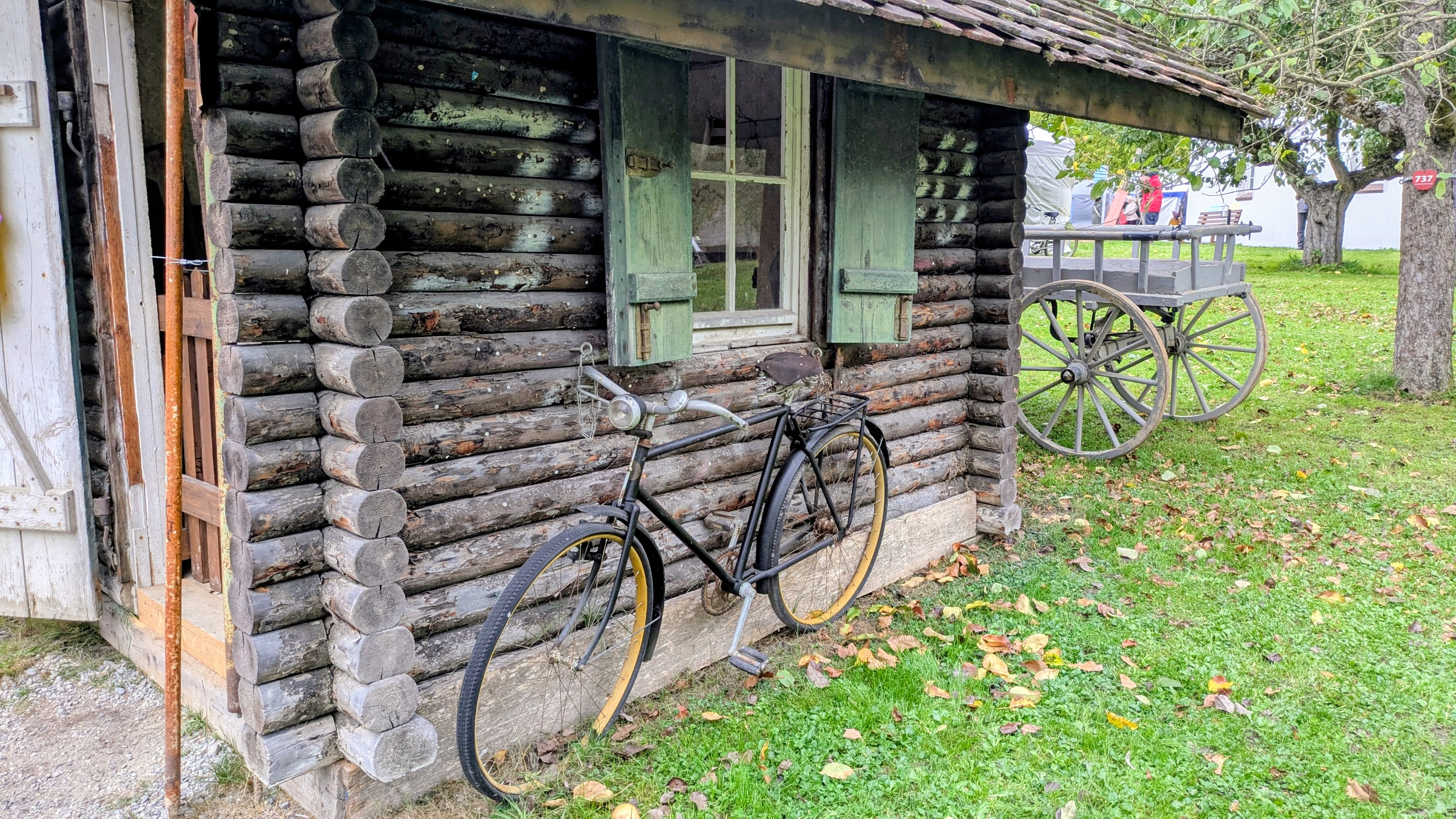 Vor einer kleinen Blockhütte aus dunklen Holzstämmen lehnt ein altes schwarzes Fahrrad mit Ledersattel und gelblichen Felgen. Die Hütte hat grün gestrichene Fensterläden und steht auf einer Wiese, auf der herbstliche Blätter verstreut liegen. Im Hintergrund ist ein grauer Holzwagen mit großen Speichenrädern unter Apfelbäumen zu sehen – eine stimmungsvolle Szene aus vergangener Zeit im Museumsdorf Kürnbach.