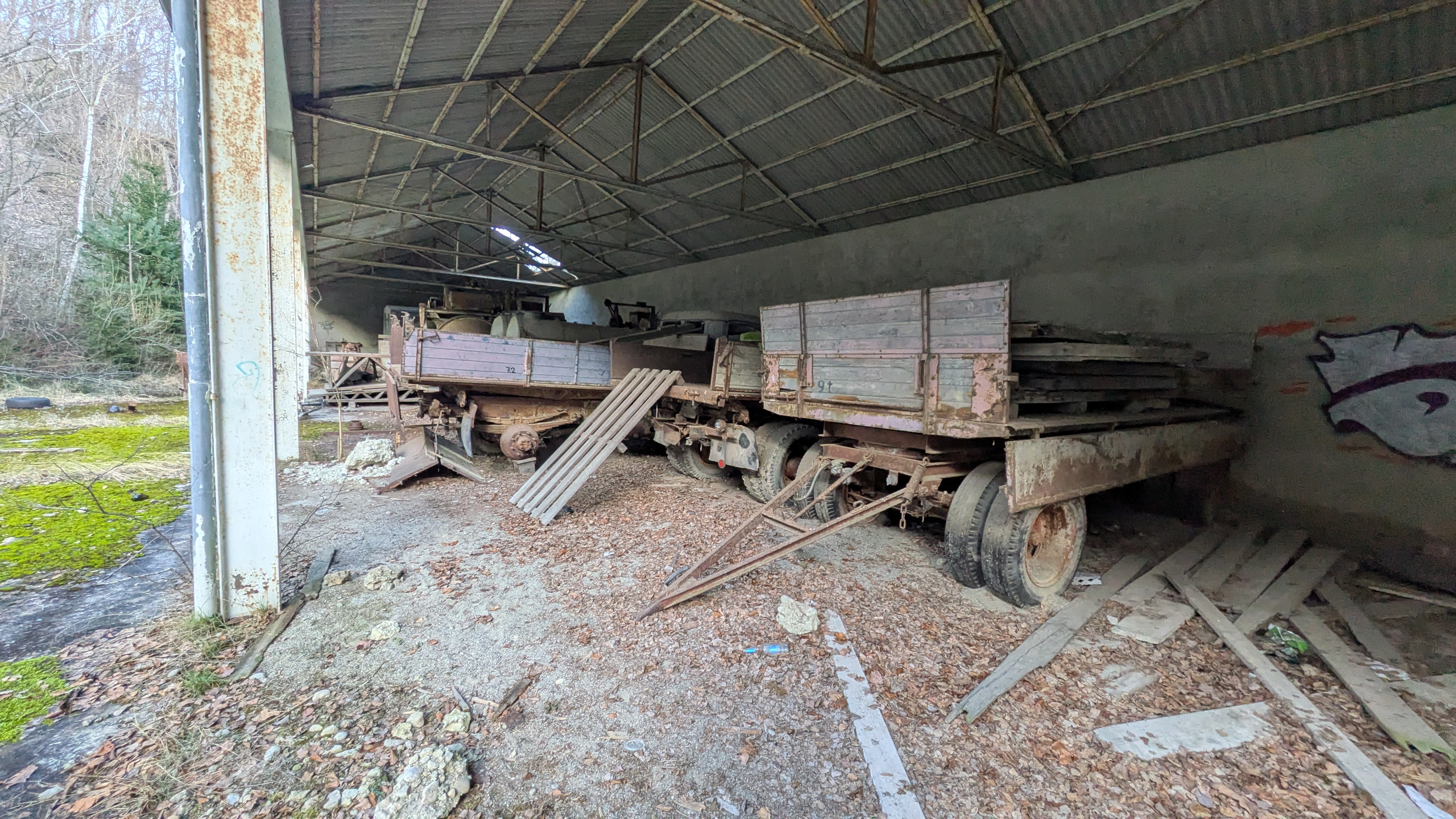 Verlassene Lagerhalle mit rostigen Anhängern und zerfallenen Holzplanken, die von der Decke durchlöchert wird