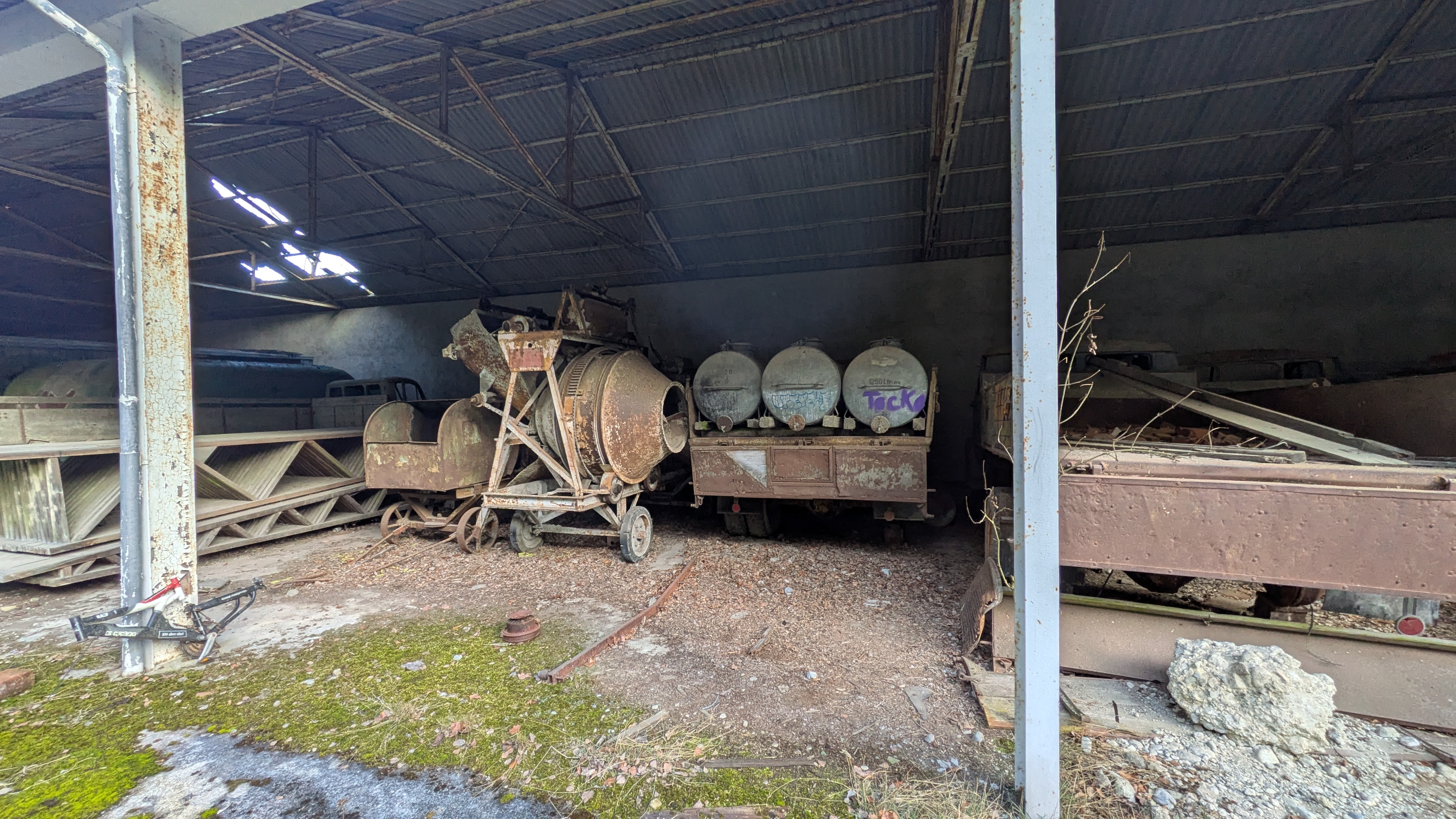 Rostige Betonmischmaschine und alte Tankwagen in einer halb eingestürzten Industriehalle.
