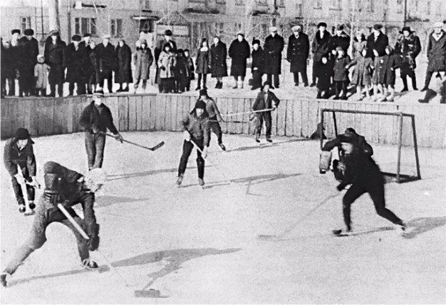 Auf einer Eisfläche spielen Männer Hockey. Viele Menschen schauen zu, meist Männer und Kinder, nur ein paar Frauen.