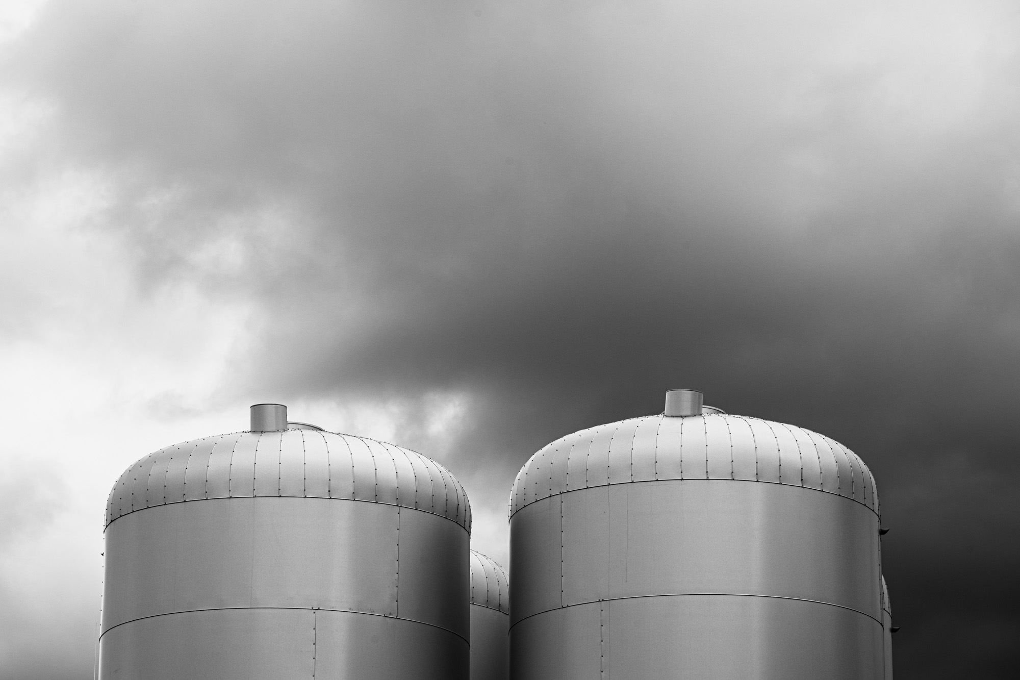 Schwarzweissbild zweier Vorratsbehälter des Blockheizwerks unseres Stadtteils. Zu sehen sind die abgeundeten Oberteile aus Stahl vor dramatisch bewölktem Himmel.