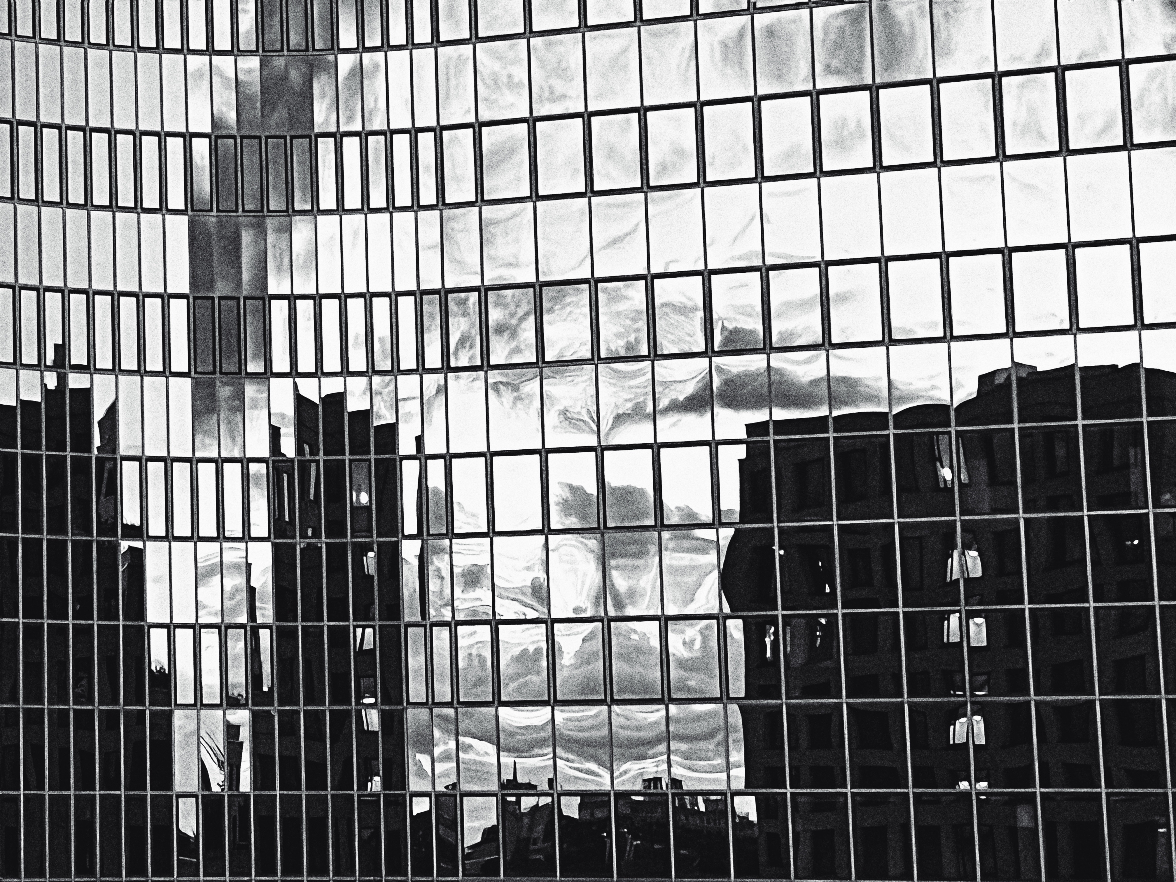 The image is a black-and-white photograph showing the reflection of a cityscape on the surface of a modern glass building. The facade consists of a grid of rectangular window panes, each slightly distorted, creating a fragmented, abstract effect.

In the reflection, several tall buildings can be seen, appearing warped and skewed by the imperfect glass. Behind them, the sky is filled with dramatic clouds, which contribute to the strong contrast and dynamic feel of the image. The interplay of light and shadow makes the clouds appear almost painted, with swirling textures and sharp divisions between dark and light.

The entire composition has a surreal, mosaic-like quality, where the reflected world looks simultaneously familiar and fragmented—evoking a sense of disorientation, depth, and urban complexity.