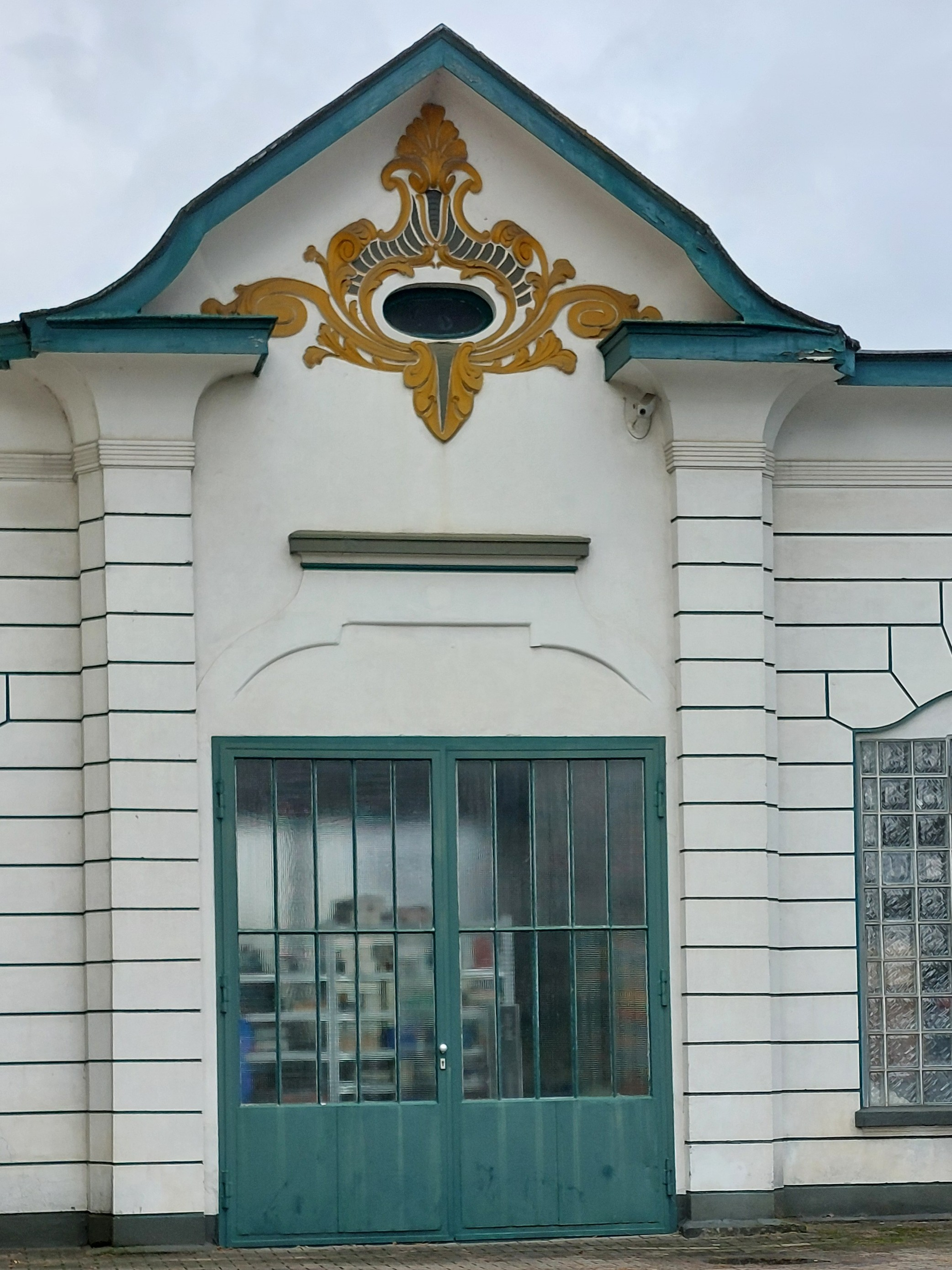 Farbfoto einer Werkstatthalle mit Stuck und Art Noveau-Elementen am Giebel verziert. Die Wände sind weiß gestrichen, das Werkstatttor mit Glaseinsätzen und die Stuckarbeiten sind in dunklem türkisgrün gestrichen, die Art Noveau Elemente sind zusätzlich in goldgelb abgesetzt.