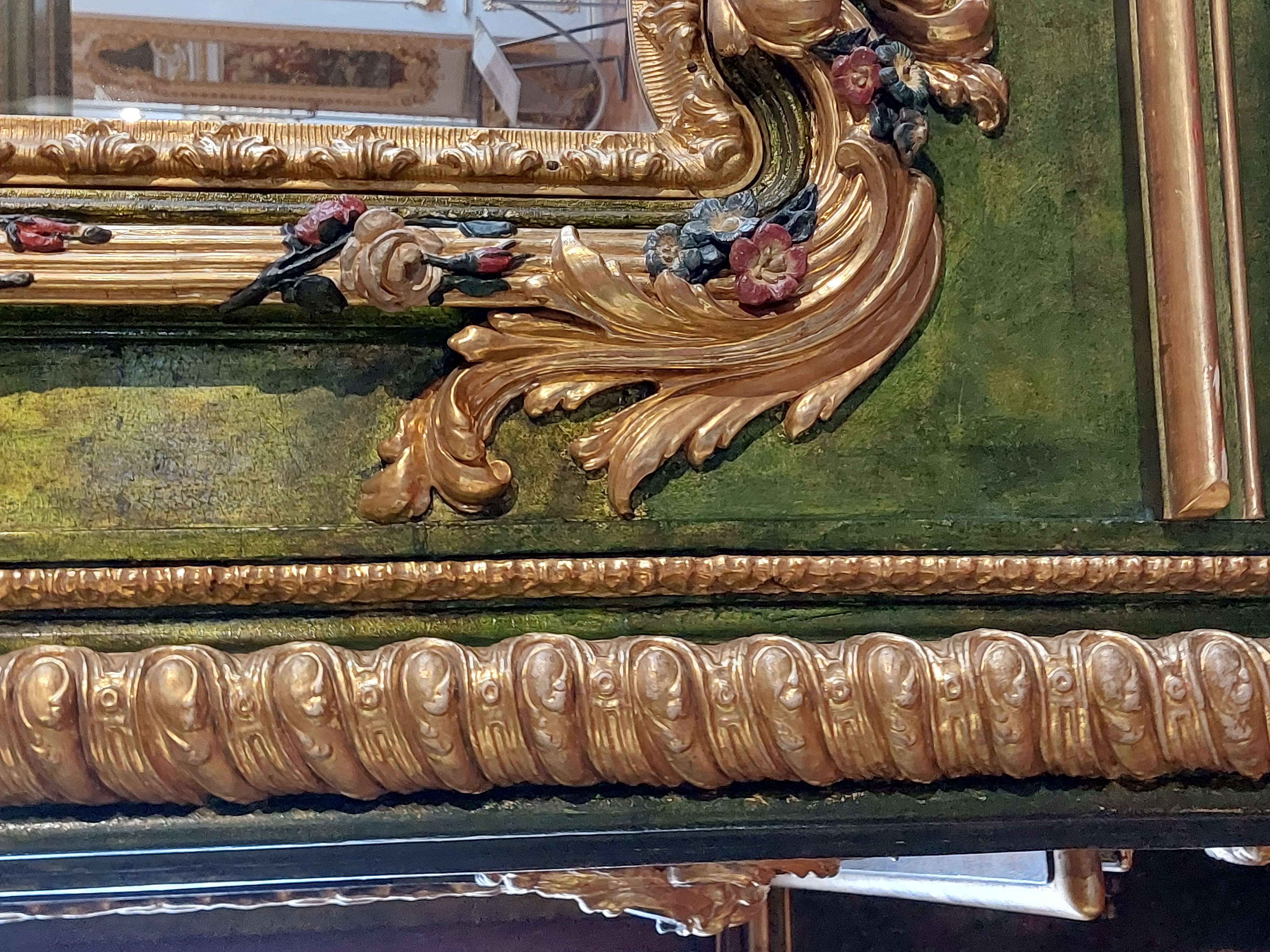 Nahaufnahme einer reich verzierten vertäfelten Wand. Die Grundfarbe der Wand ist ein leicht fleckiges gelbliches grün, die floralen Schmuckelemente sind in hellem gelbgold mit einzelnen Farbakzenten gehalten, am Rand ist ein eingefasster Spiegel sichtbar.