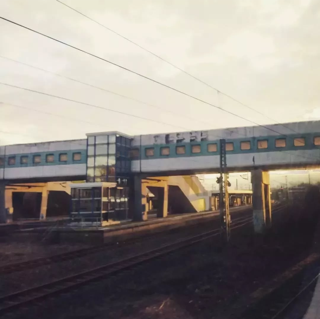 Gelbstichiges Foto einer überbauten Brücke über Eisenbahngleise mit dahinter sichtbarem Inselbahnsteig. Die Überbauung der Fußgängerbrücke besteht aus gerundeten Betonelementen mit eingesetzten quadratischen Fenstern mit abgerundeten Ecken. Der Überbau ist wie ein Zug gestrichen, hellgrau mit durchgehend blauem breitem Streifen entlang der Fenster.

Angebaut ist ein in Glas eingehauster Aufzug, in dem sich die Umgebung spiegelt. 

Die aufgenommene Szene wird von tiefstehender Sonne bestrahlt und wirkt deswegen gelblich.
