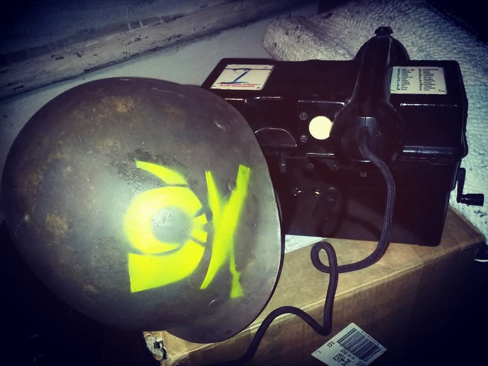 Leicht überbelichtetes Foto eines schwarzen, kastigen Feldtelefons und einem gräulich lackierten Stahlhelm mit Rostflecken und gelb aufgesprühtem Posthorn.