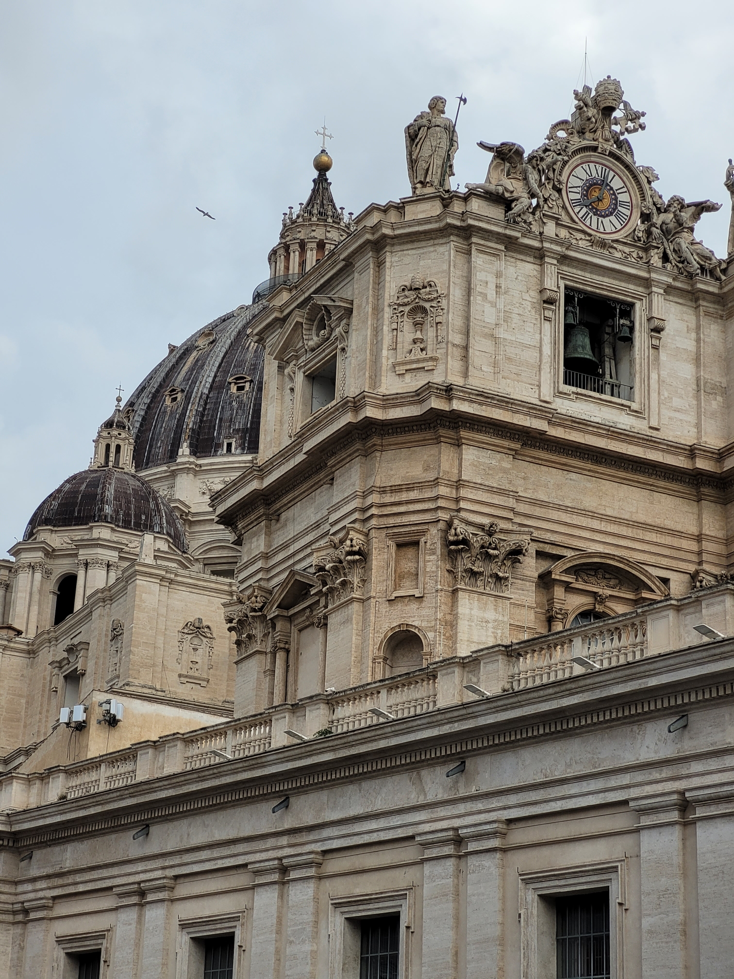 parts of Saint-Peter's basilica seen from the south side: the southern top angle of the façade, the huge dome behind it, and a smaller dome on the side. on the lower part of the picture is the southern arm that embraces Saint-Peter's square. cloudy sky and a bird.