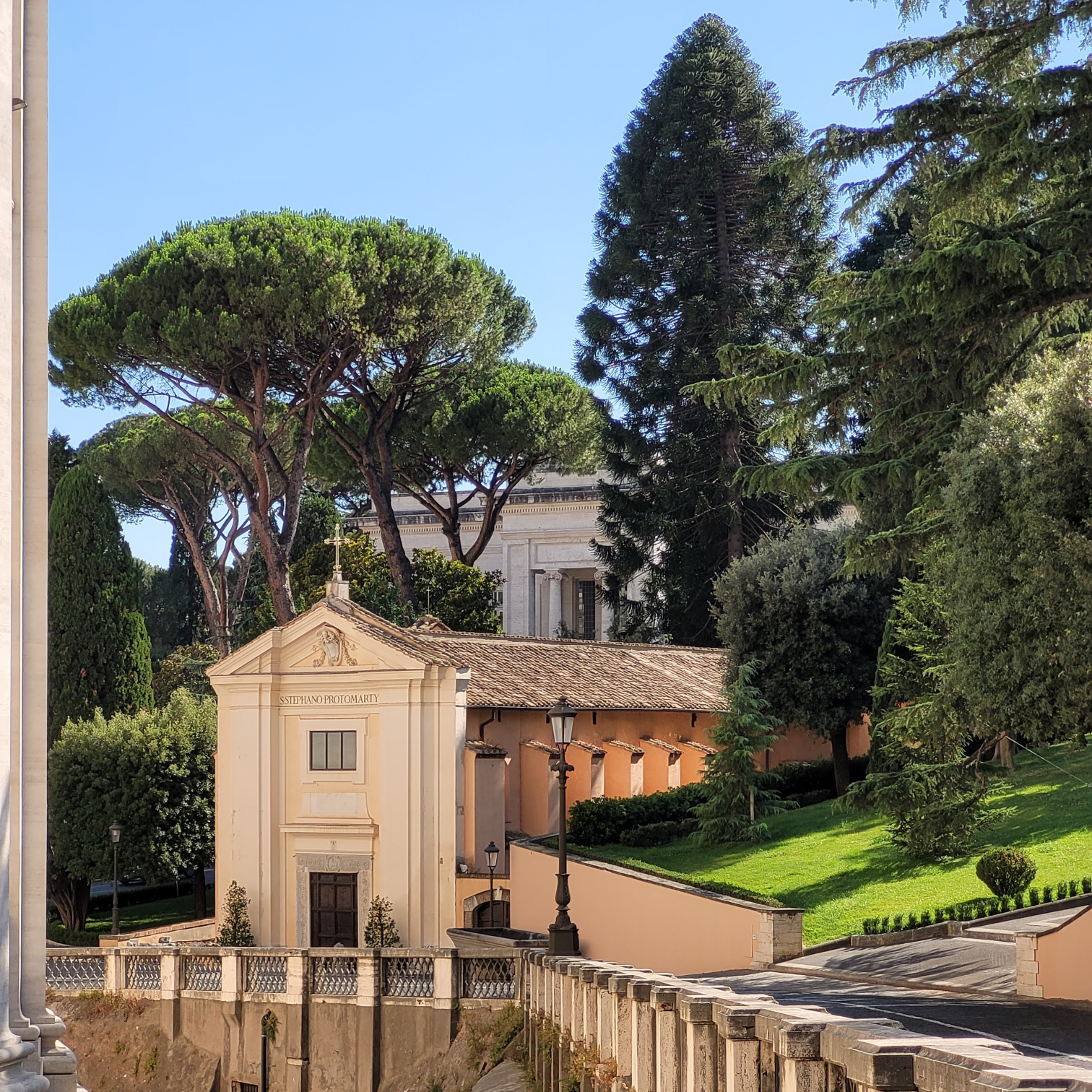 a square picture. in the center, the front of the little church of Saint-Stephen. far behind it, the front of the Vatican's railway station. on the left, the high walls of the back apse of Saint-Peter's. trees.