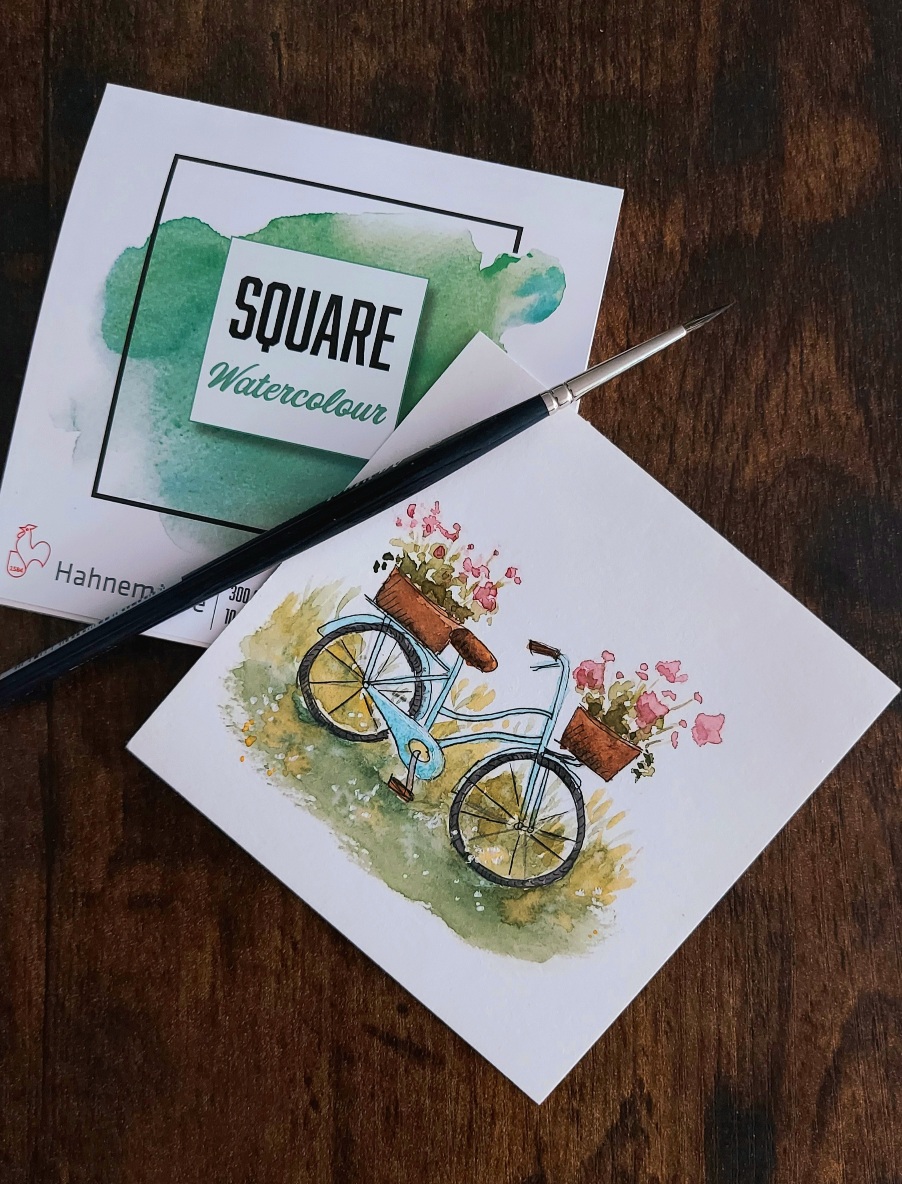 Kleines quadratisches Aquarell mit einem türkisblauen Fahrrad auf einer Wiese. Vorne und hinten sind Blumenkörbe auf dem Rad.