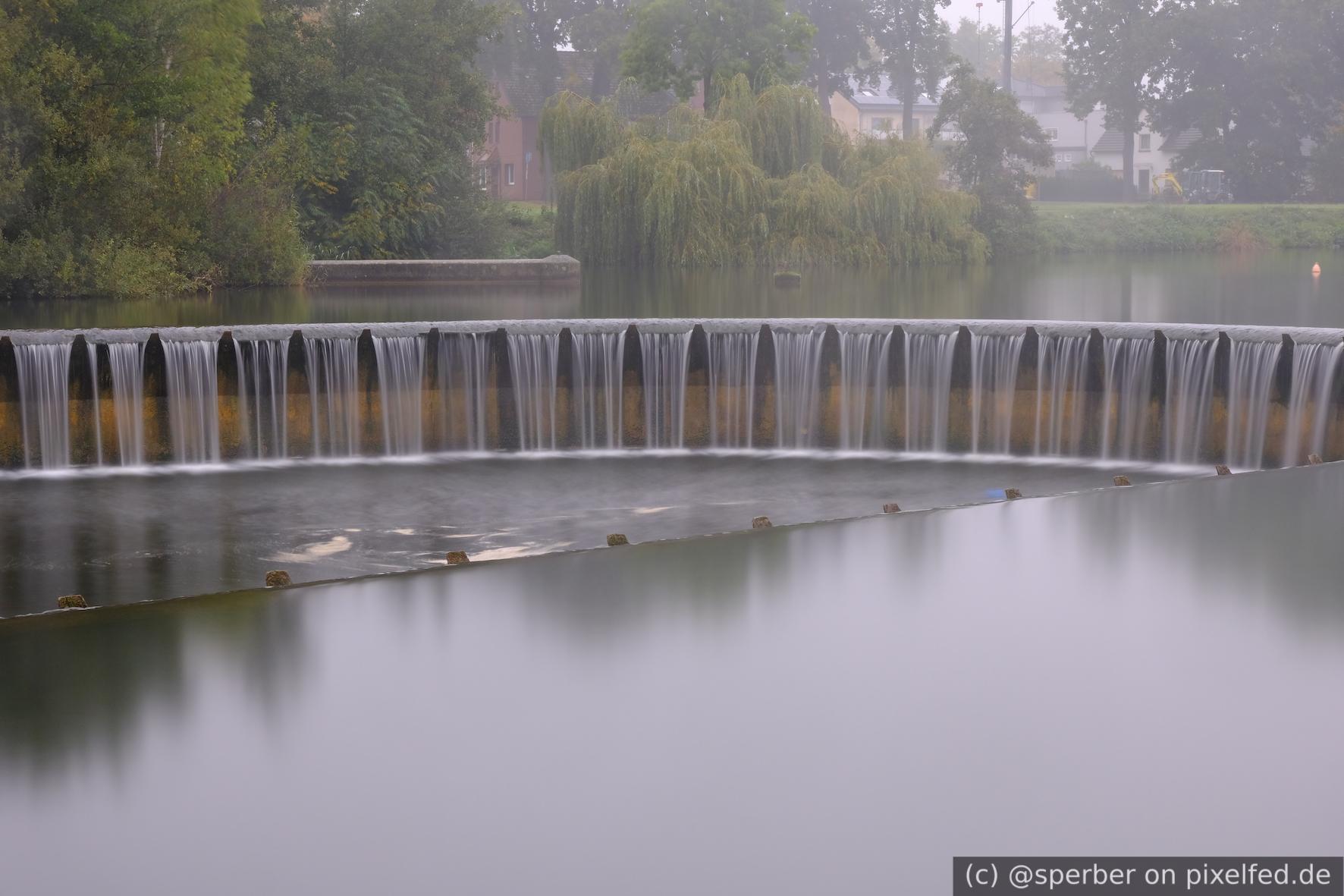 Round overflow of a lake with park in the background. The water looks smooth, due to the long exposure time.