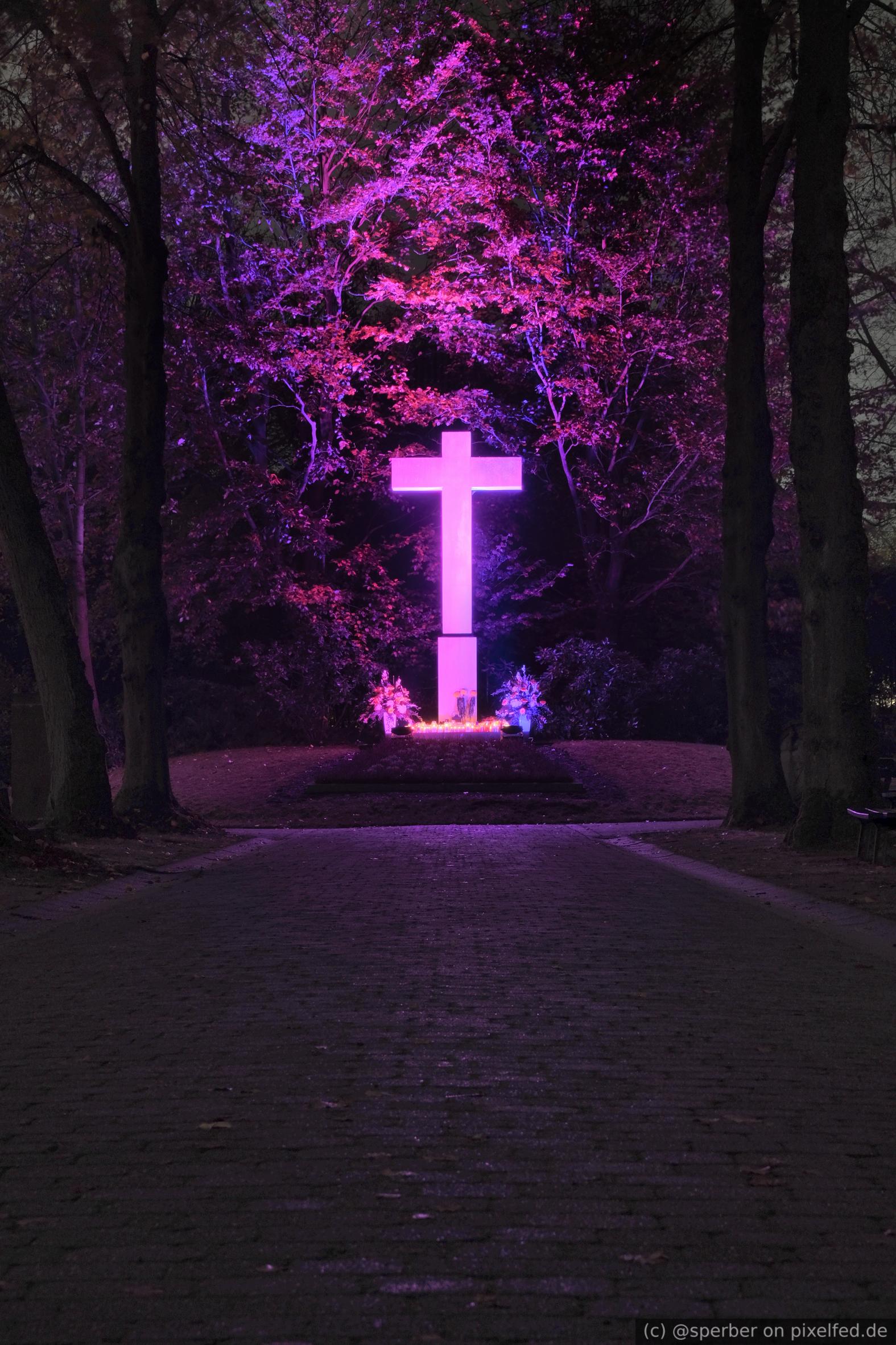 A dark alley leading to a white cross with purple illumination and candles.