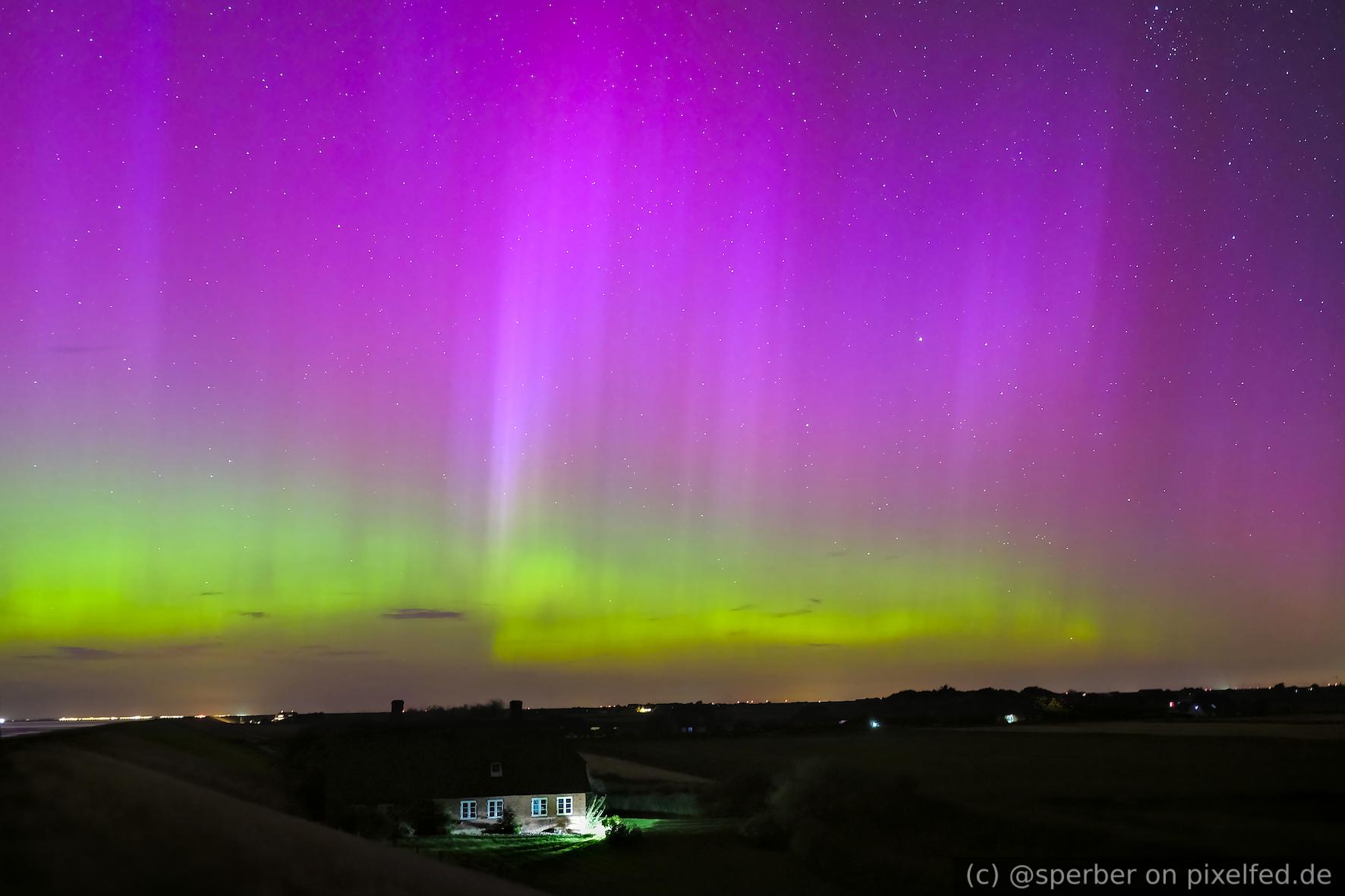 Single illuminated house with the island dike on the left and fields on the right. The sky is illuminated by a green and purple aurora.