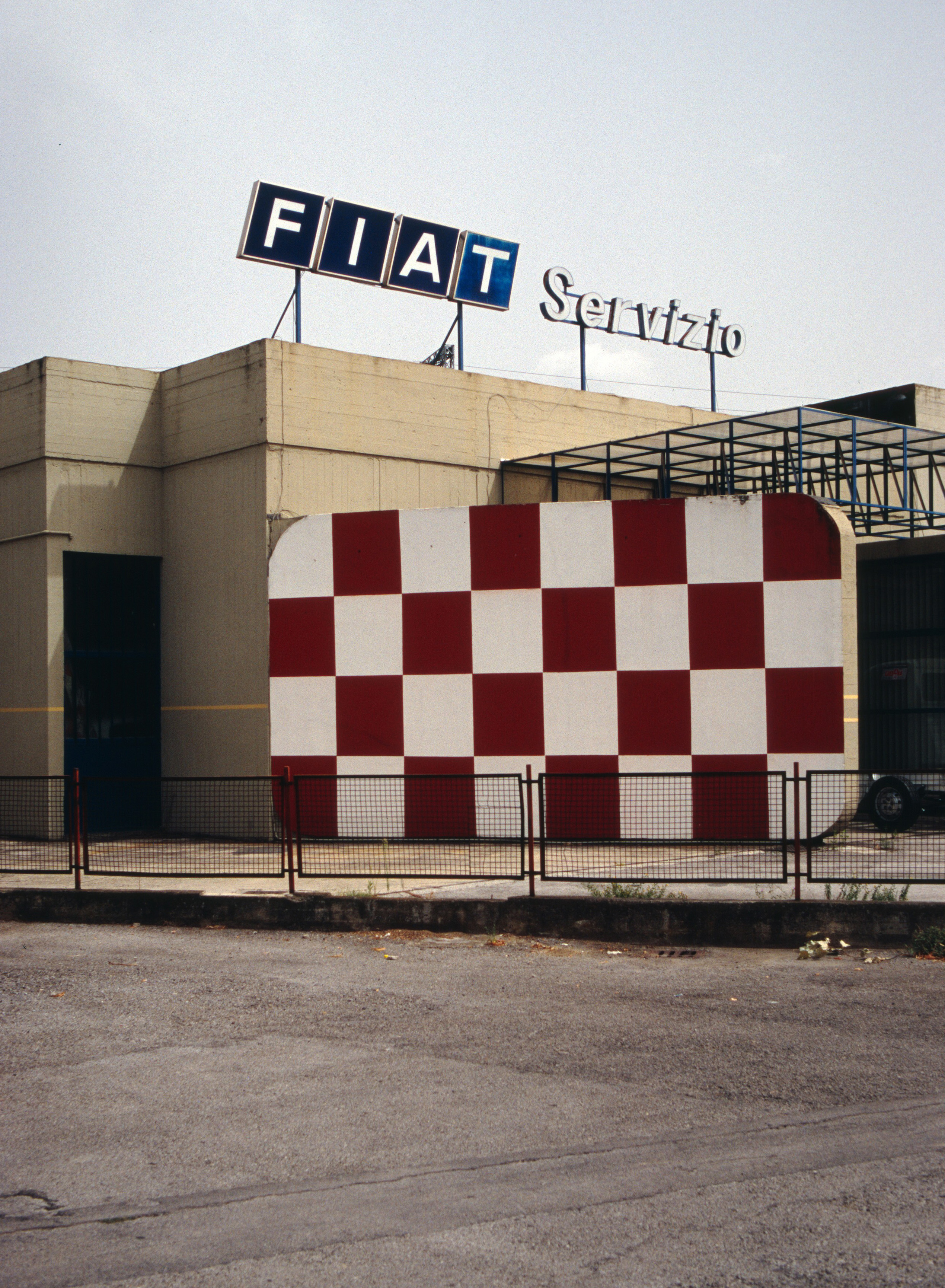 Foto eine Autowerkstatt in Italien. Der Schriftzug "Fiat Servizio" steht auf dem Dach darunter eine Mauer mit auffälligem rot-weißem Karo-Muster.