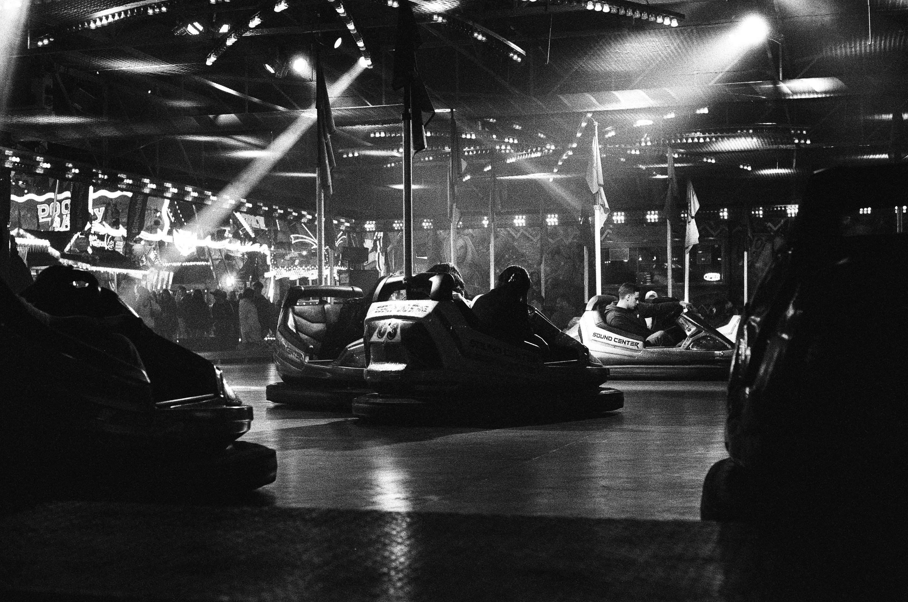 A grainy black and white photograph of a bumper car area. Light beams are visibly illuminating parts of the area, in the background more fair attractions are visible.