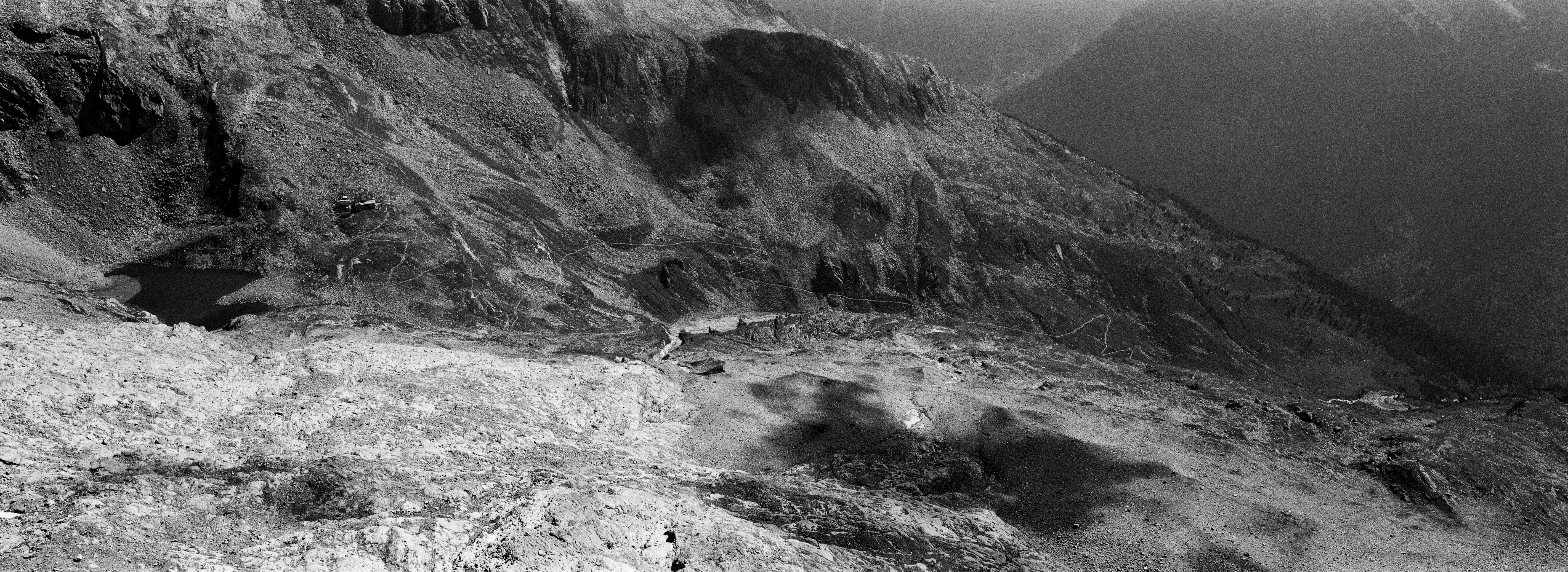 A panoramic landscape photo in black-and-white. It shows a hiking path sneaking up along the side of a mountain, all the way from the right to left of the entire picture