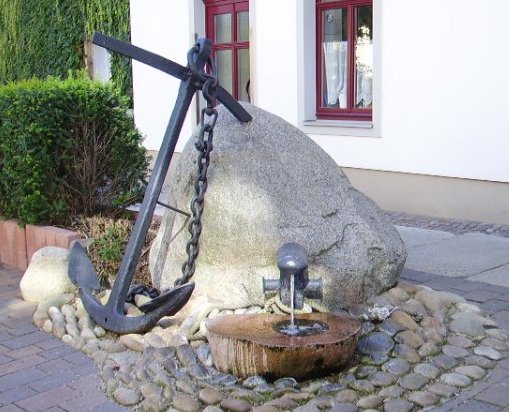 Gepflasterter Platz mit Quellstein und angelehntem Anker