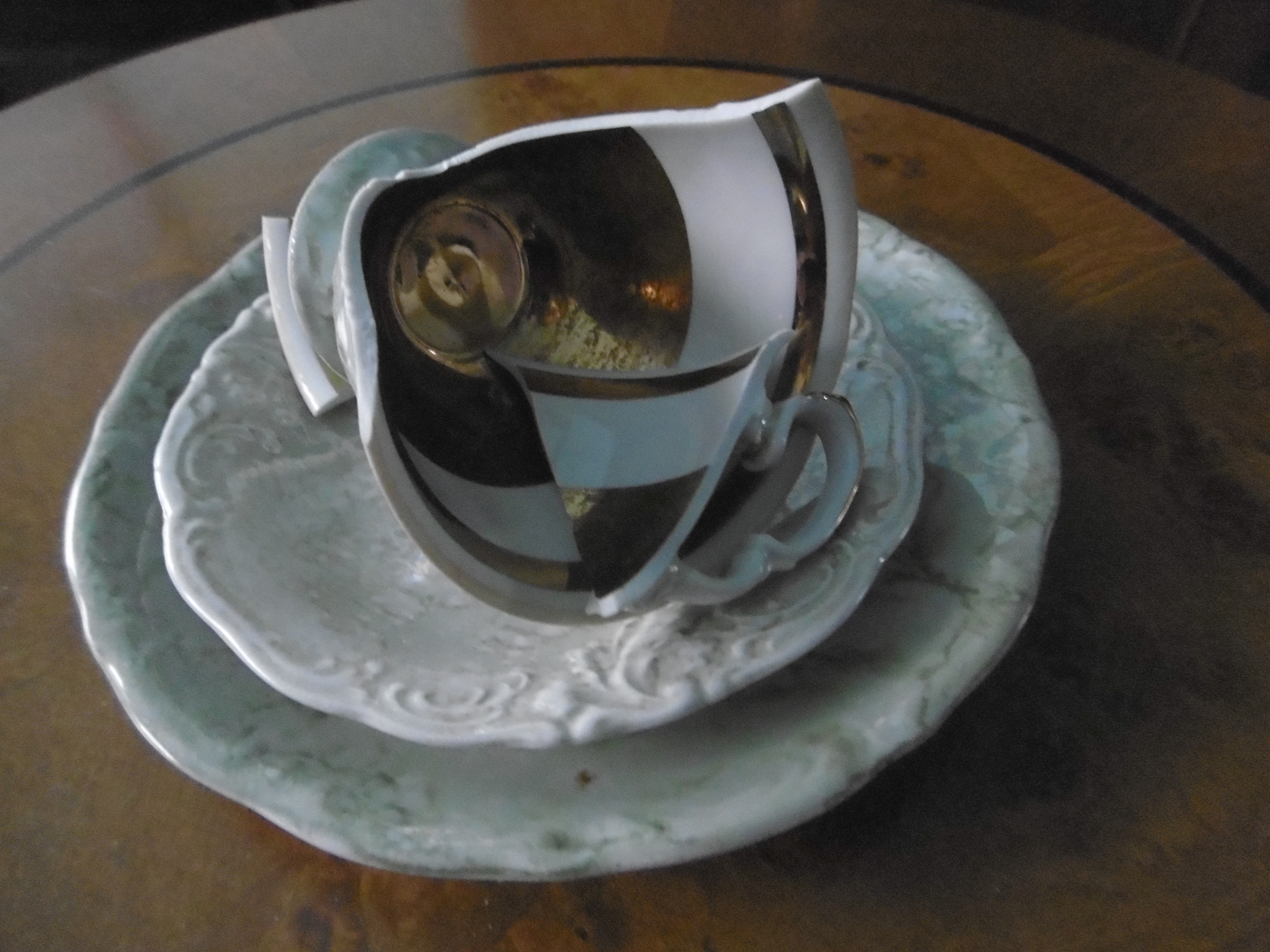 Auf einem Tisch steht ein Teller, darauf eine Untertasse und darauf liegt eine zerbrochene Tasse, das Gedeck ist grün und gold