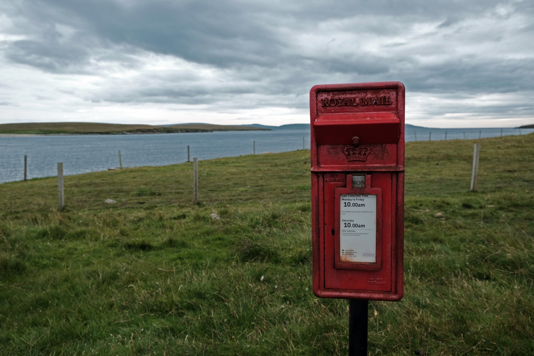 A red mailbox stands in front of a green field bordering a sound. The sky is covered with dark clouds