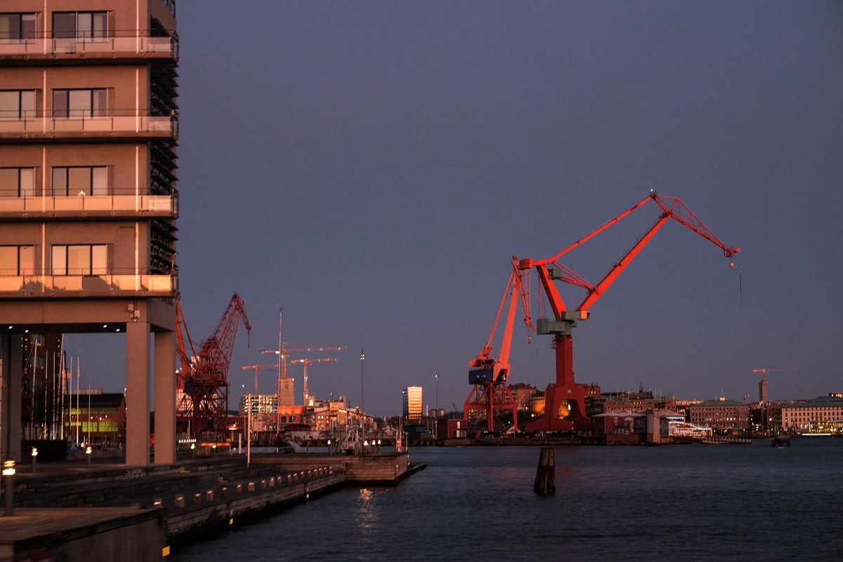 Direkt am Rand eines Gewässers steht rechts ein schmales Hochhaus auf Stelzen, so dass an entlang einer Kaimauer Fahrzeuge unter dem Gebäude hindurchfahren können. Das Gebäude ist in goldenes Abendlicht getaucht, der Himmel dahinter ist bereits dunkel. Davor sind noch mehrere rote Hafenkräne zu sehen.