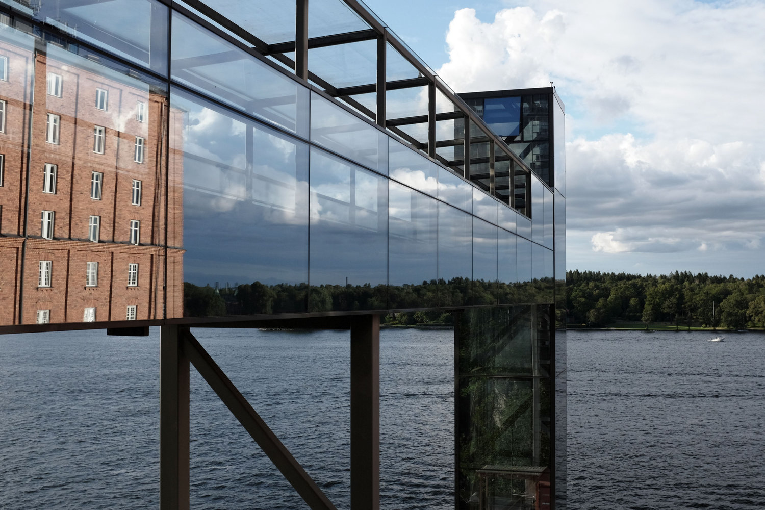 Von links ragt eine Glas-Stahl-Konstruktion zunächst waagerecht ins Bild. An ihrem Ende führt ein senkrechter Schacht nach unten. Dahinter sind Wasser und eine mit Bäumen bewachsene Uferlinie zu sehen. In den Glaswänden des waagerechten Konstruktionsteils spiegeln sich Teile der gegenüberliegenden Uferlinie, einige Wolken sowie ein rotes Backsteingebäude.