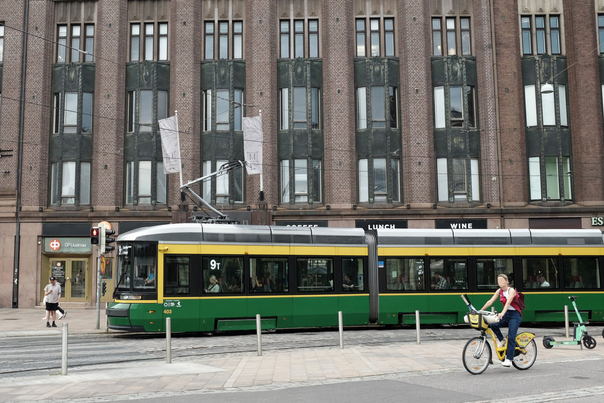 Vor einem Büro- und Geschäftsgebäude aus Backstein fährt eine grün-gelb-lackierte Straßenbahn von rechts ins Bild. Parallel zur Straßenbahn auf einem extra Radweg fährt eine Frau auf einem gelben Fahrrad. An einer Ampel steht eine Person mit Baseballmütze.