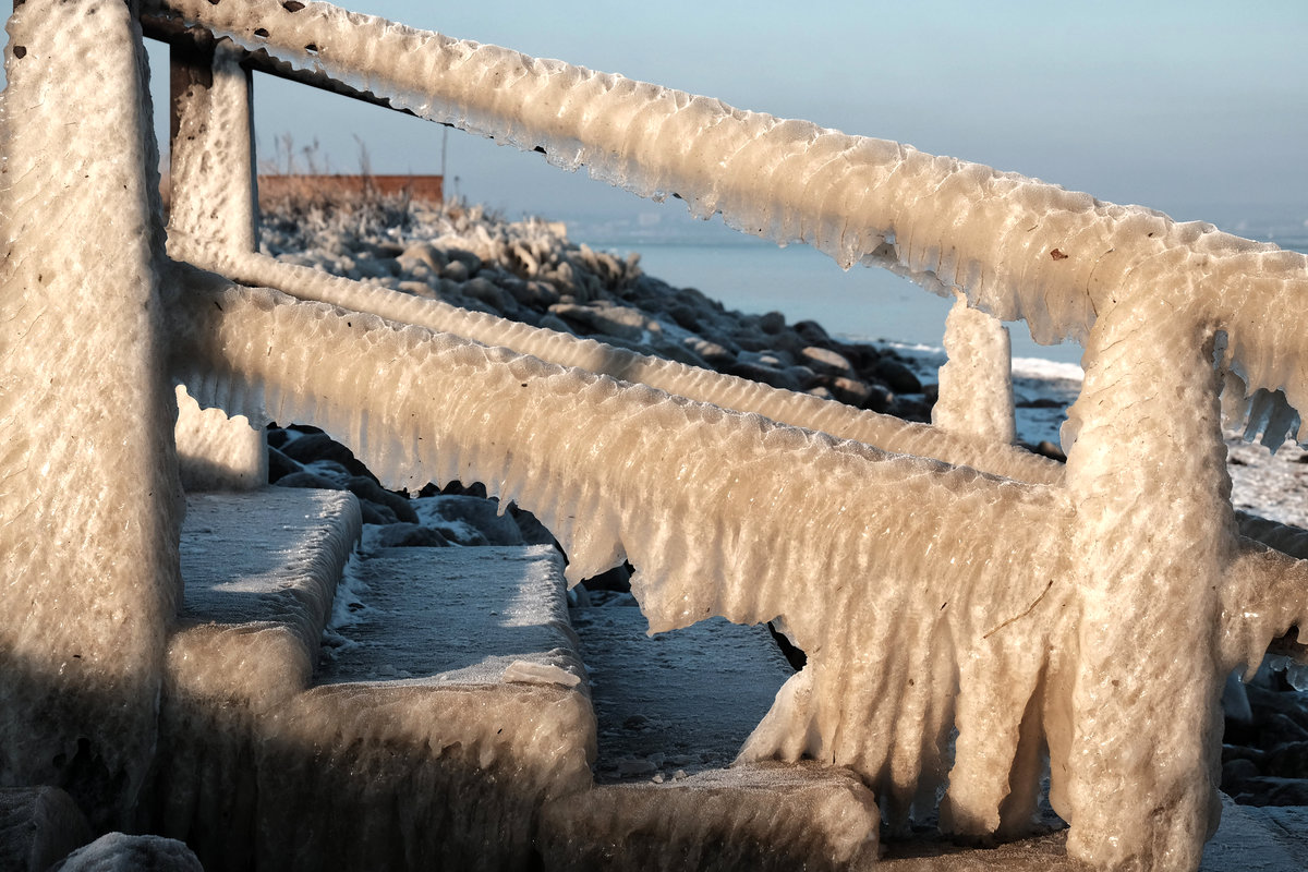 Eine flache Holztreppe an einem Strand seitlich aufgenommen. Die Stufen sind dick vereist, vom Treppengeländer hängen Eismassen, die vom Wind so geformt sind, dass sie flatternden Stoffstücken ähneln