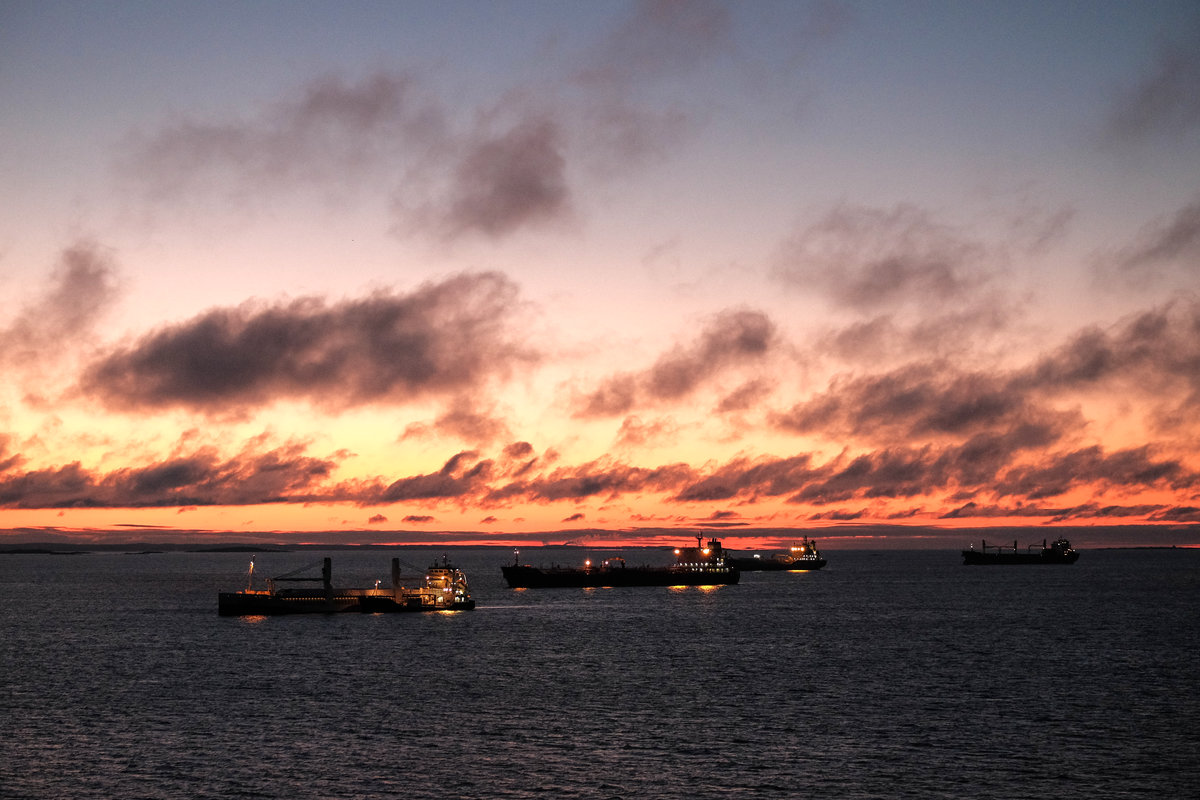 Drei Schiffe liegen vor einigen flachen Felseninseln vor Anker. Der Himmel über dem Horizont färbt sich golden, sonst herrscht noch Dunkelheit. Oberdecksbeleuchtung und Ankerlichter erhellen die Schiffe ein wenig.