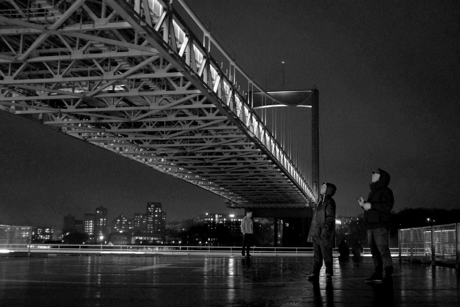 Schwarzweiß-Bild eines glatten, spiegelnden Schiffsdecks bei Dunkelheit. Zwei Personen stehen auf dem Deck und blicken nach oben zu einer erleuchteten Hängebrücke, die von links oben nach rechts unten durchs Bild verläuft. Sie erscheint für die Personen auf dem Deck sehr nah.