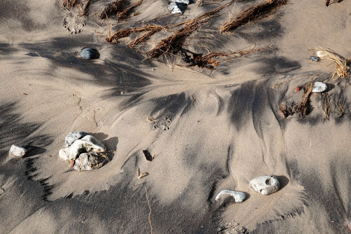 Heller und dunkel gefärbter Sand bilden Muster, die aussehen wie ineinander verlaufende Farben. Dazwischen liegen einige helle Steine und Seetang.