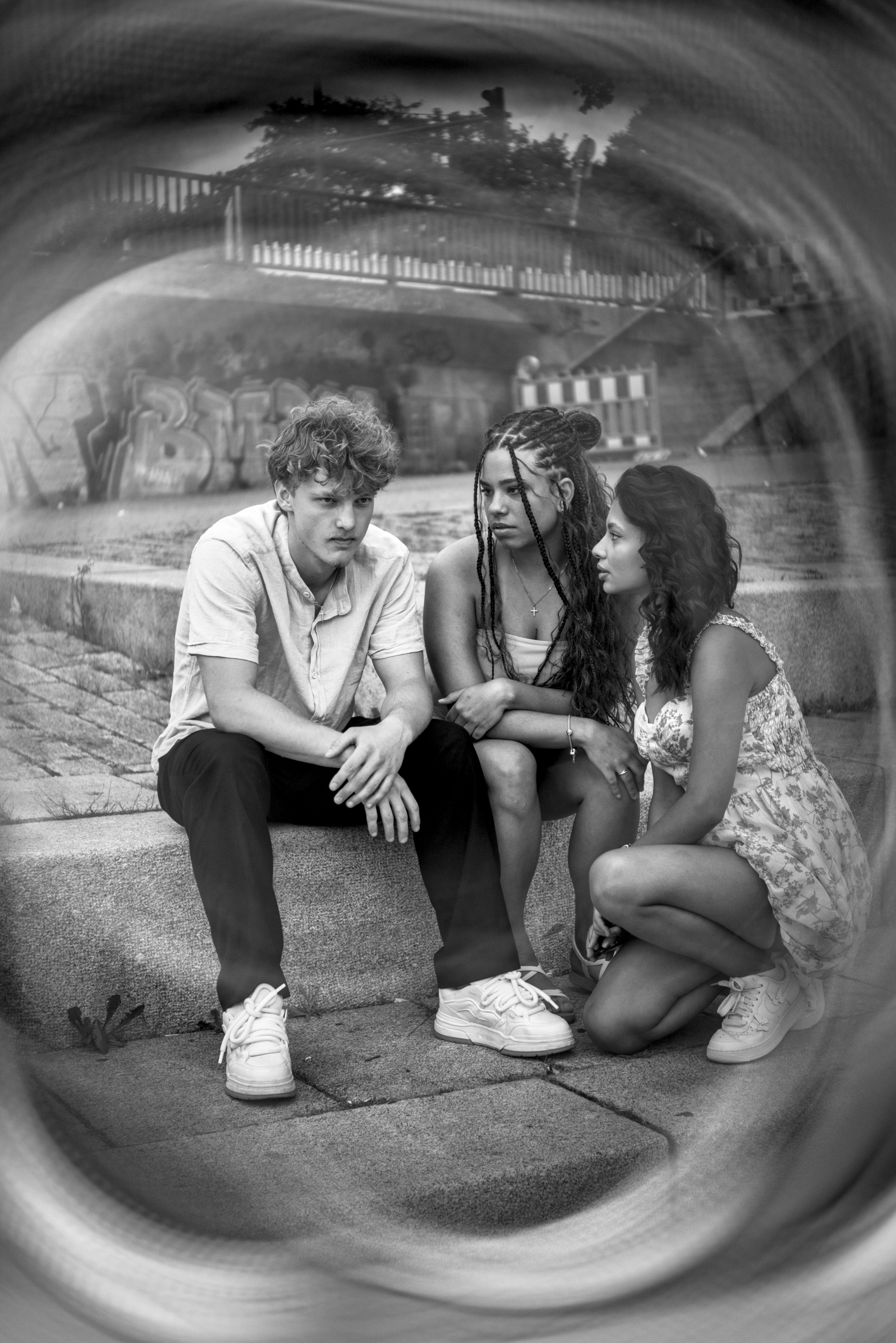 Ein Schwarzweißfoto. Ein männlich gelesenes Model und zwei weiblich gelesene Models sitzen auf einer großen Stufe. Der Rand des Fotos ist verzerrt von einer Effekt-Linse.
