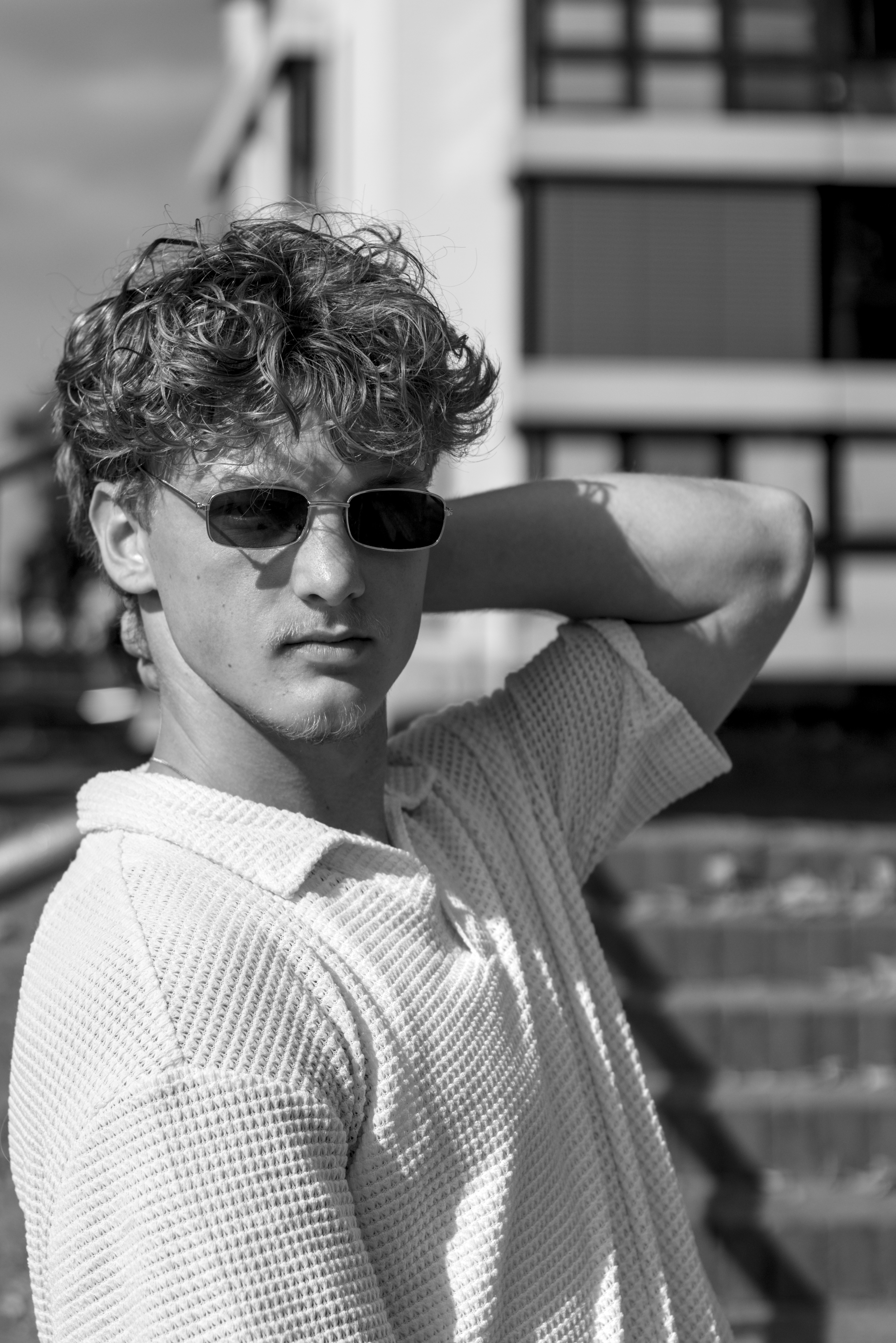 Ein Schwarzweißfoto. Ein männlich gelesenes Model auf einer Treppe. Er trägt ein weißes Shirt und eine Sonnenbrille.