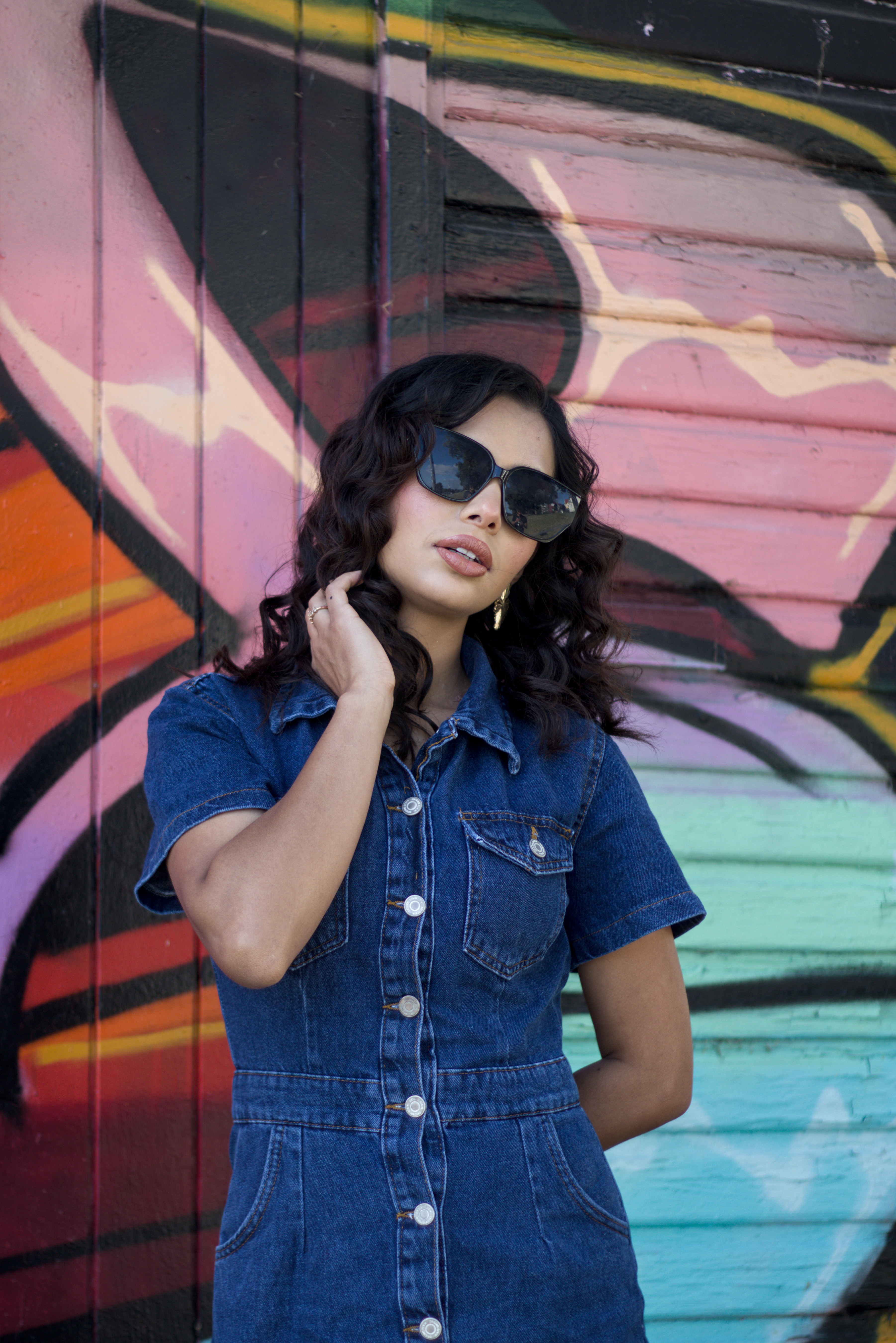 Ein Farbfoto. Ein weiblich gelesenes Model vor einer bunten Holzwand. Sie hat schwarze gelockte Haare und trägt ein blaues Jeanskleid und eine Sonnenbrille.