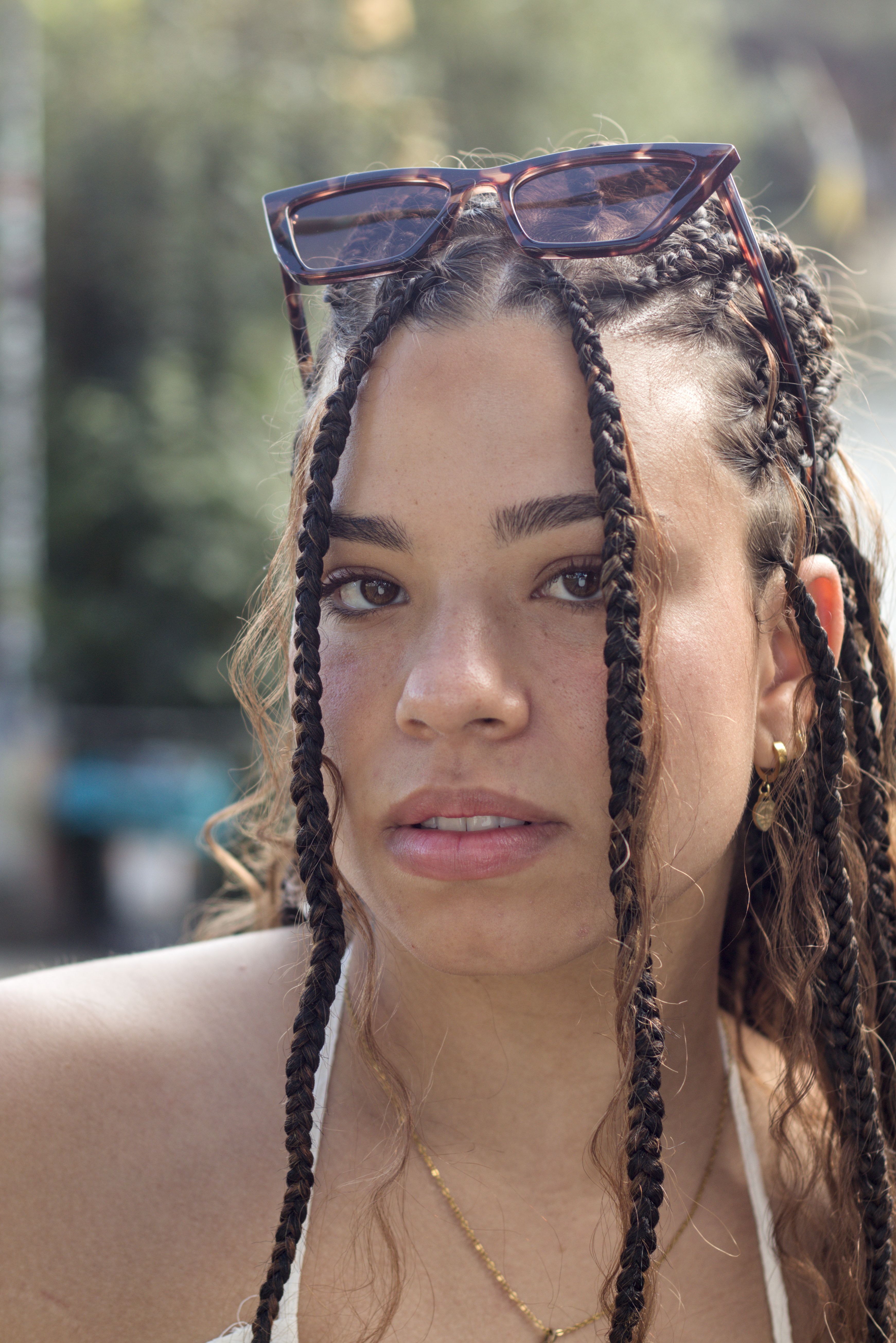 Ein Farbfoto. Ein weiblich gelesenes Model mit geflochtenen Haaren und einer Sonnerbrille auf dem Kopf.