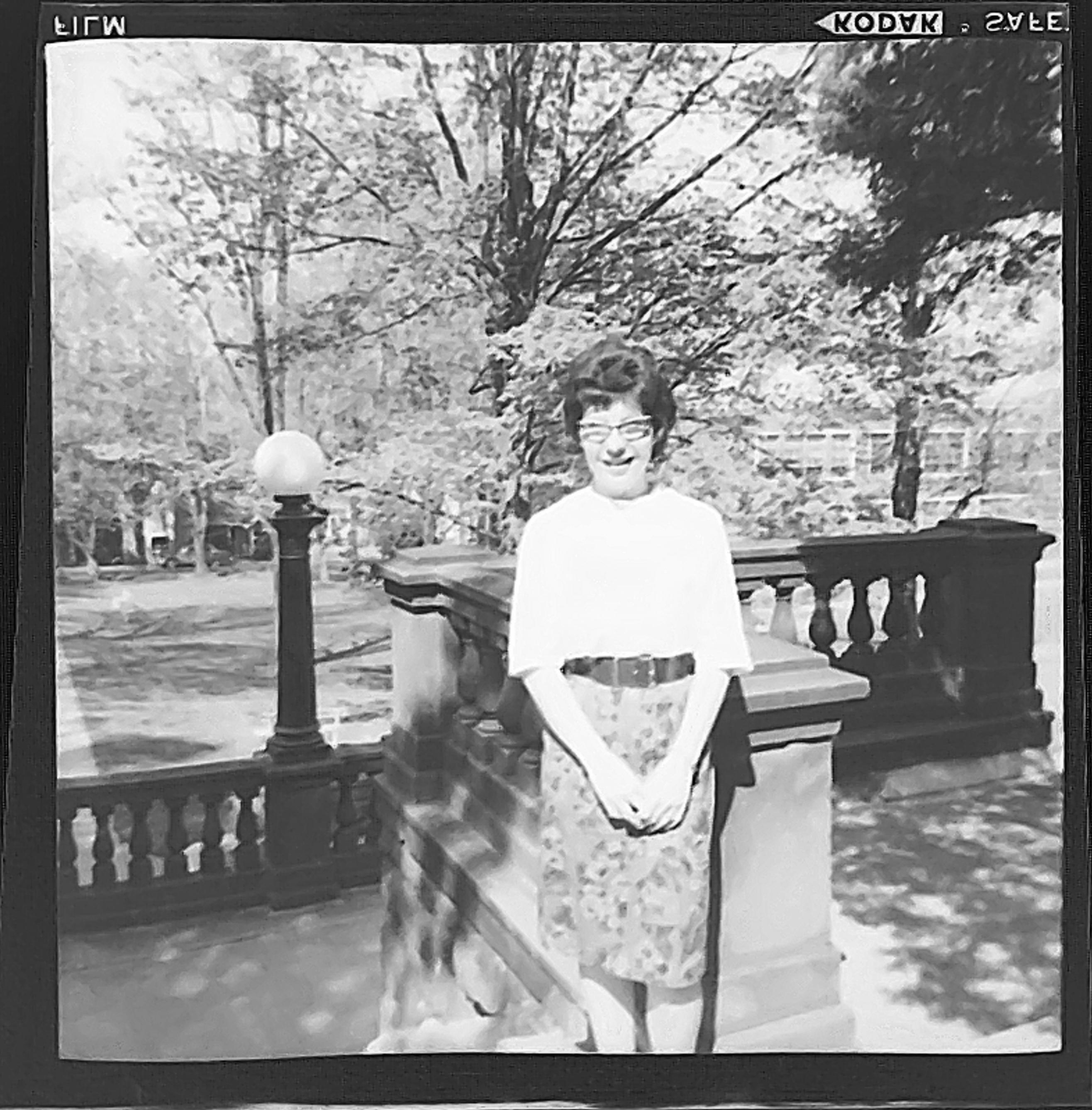 Black and white shot of a woman posing for the camera in a park. We're at the top of some stone balustrades. There's a lamppost with a globe on top.