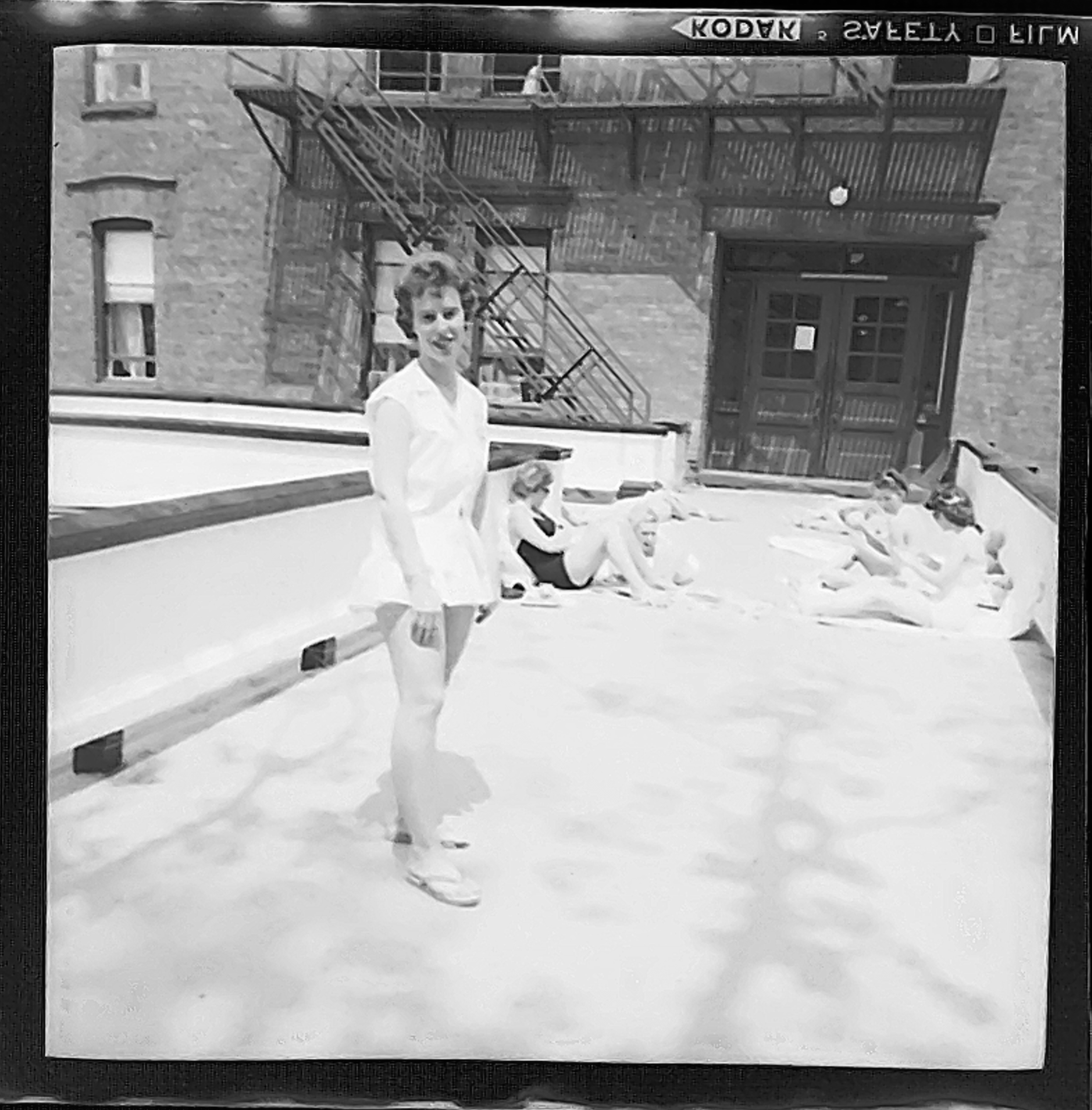 Black and white shot of a young woman standing on a roof where some other young women are sunning themselves. They're all wearing modest, one-piece swimsuits.