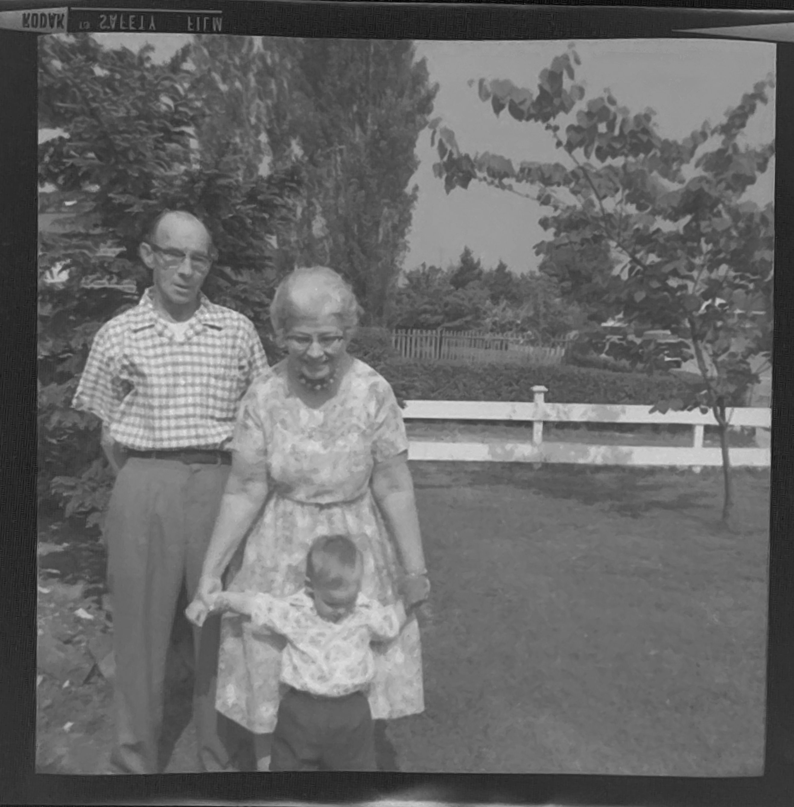 A white-haired woman is holding out the arms of a young boy. Behind her is a rather serious-looking gentleman. Black and white photo.