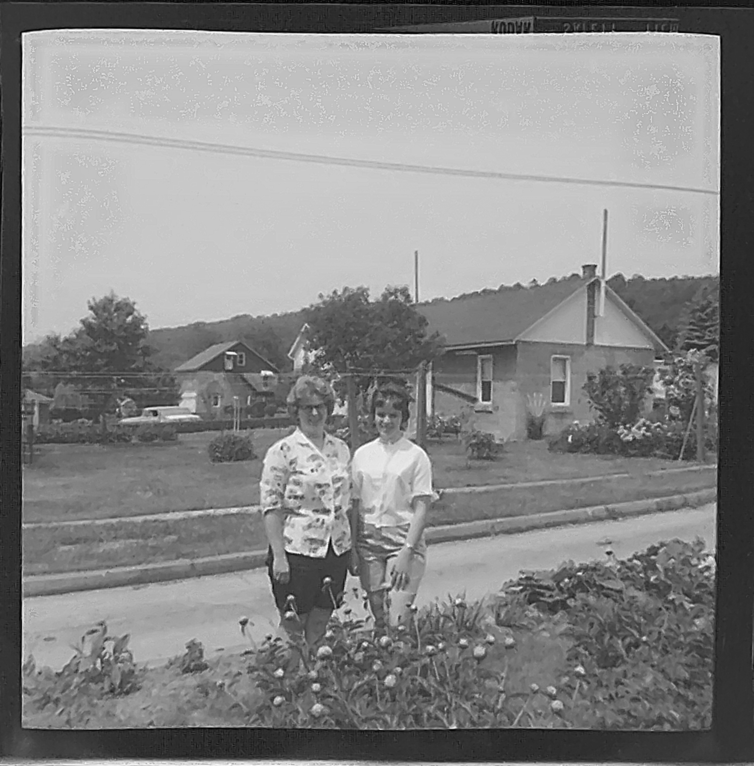 Black and white shot of two women. Looks like the shot was taken in a front yard, looking over some low flower beds.