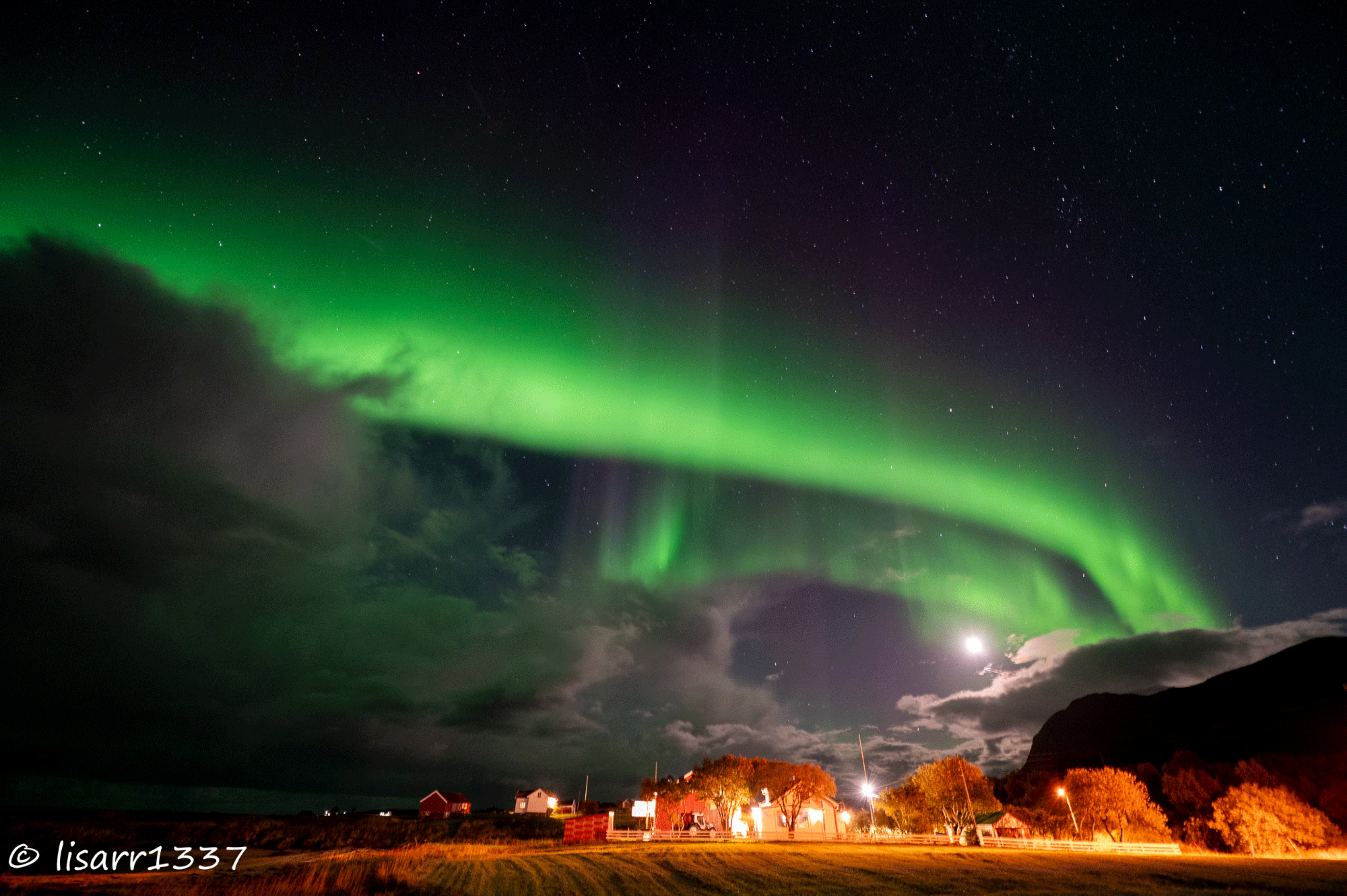 GIF mit Zeitreihe einer Aurora über ein paar beleuchteten Häusern bei Vollmond.