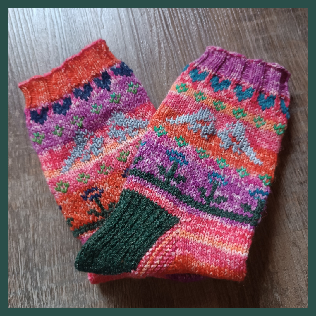 A dark green frame surrounds the picture of a pair of handknit socks. They are folded at the heel to showcase the leg with different colourwork motifs. The base colour is orange and purple with a dark green heel. The colourwork motifs are from top to bottom: blue hearts, green blossoms, grey love birds, green blossoms, blue and green flowers standing on a strip of grass.