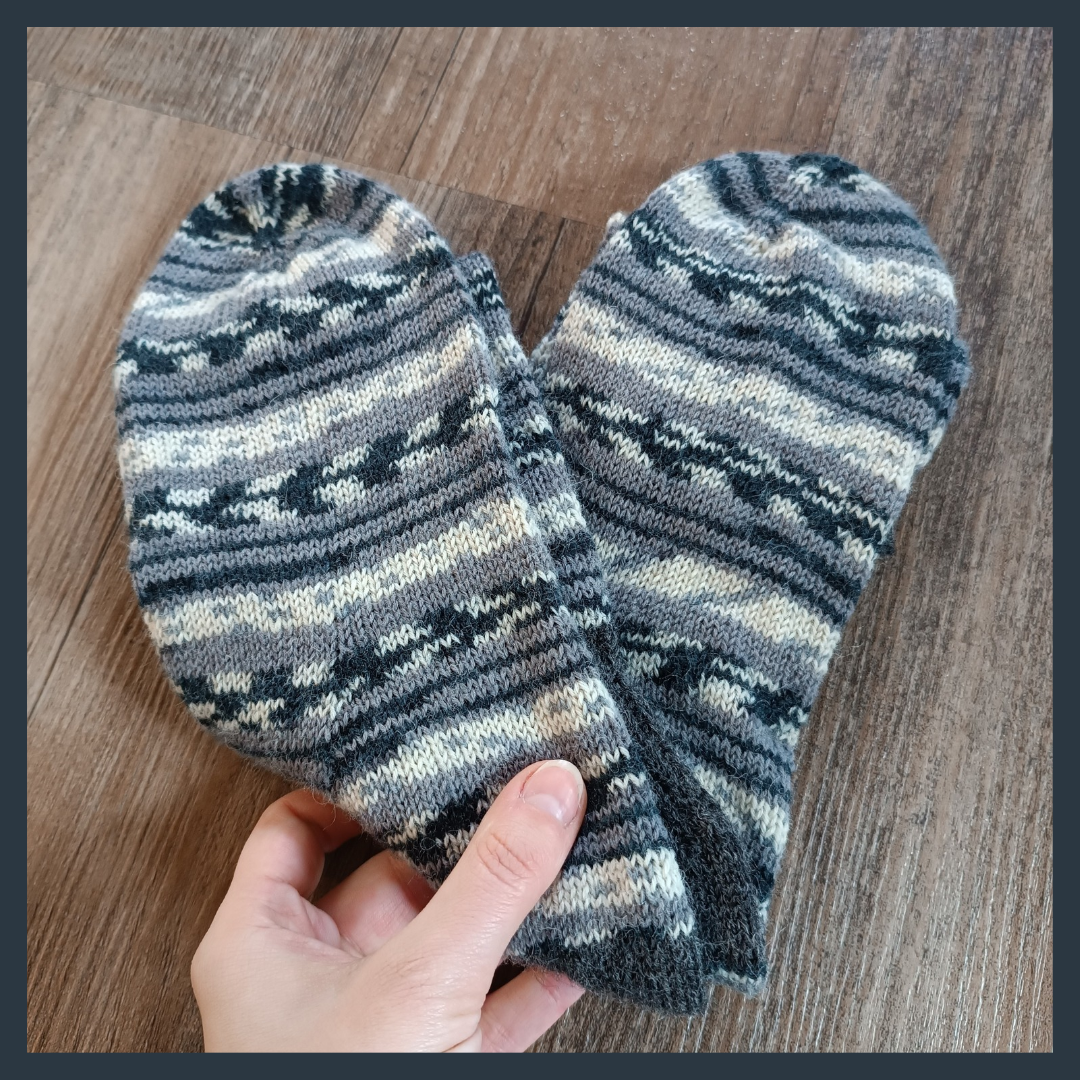 A dark grey frame surrounds the picture of a pair of handknit socks on a wooden floor. A light skinned hand holds them from the bottom of the picture. The socks are folded into a heart shape. They are made of a self-striping yarn in white, grey and black. The stripes show different patterns like dots, smaller stripes or arrow shapes.