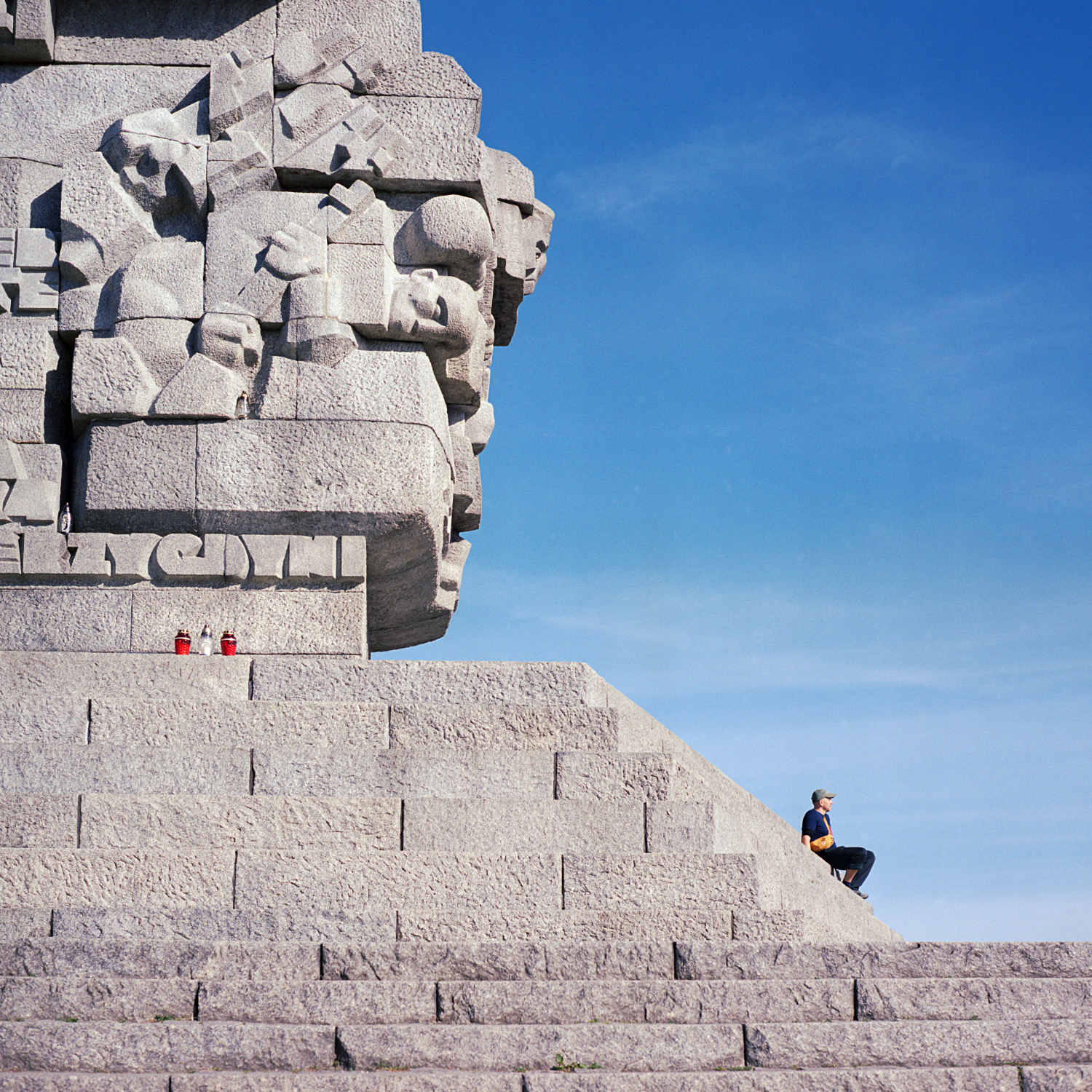 Person sitting on wide stone steps of a large monument with carved reliefs, five candles placed above, under a clear blue sky.