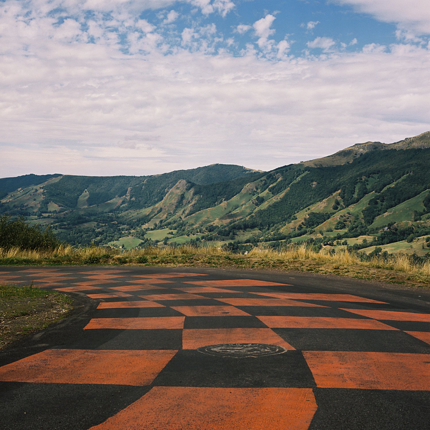 Checkered orange and grey road, overlooking rolling green mountains under a partly cloudy sky.