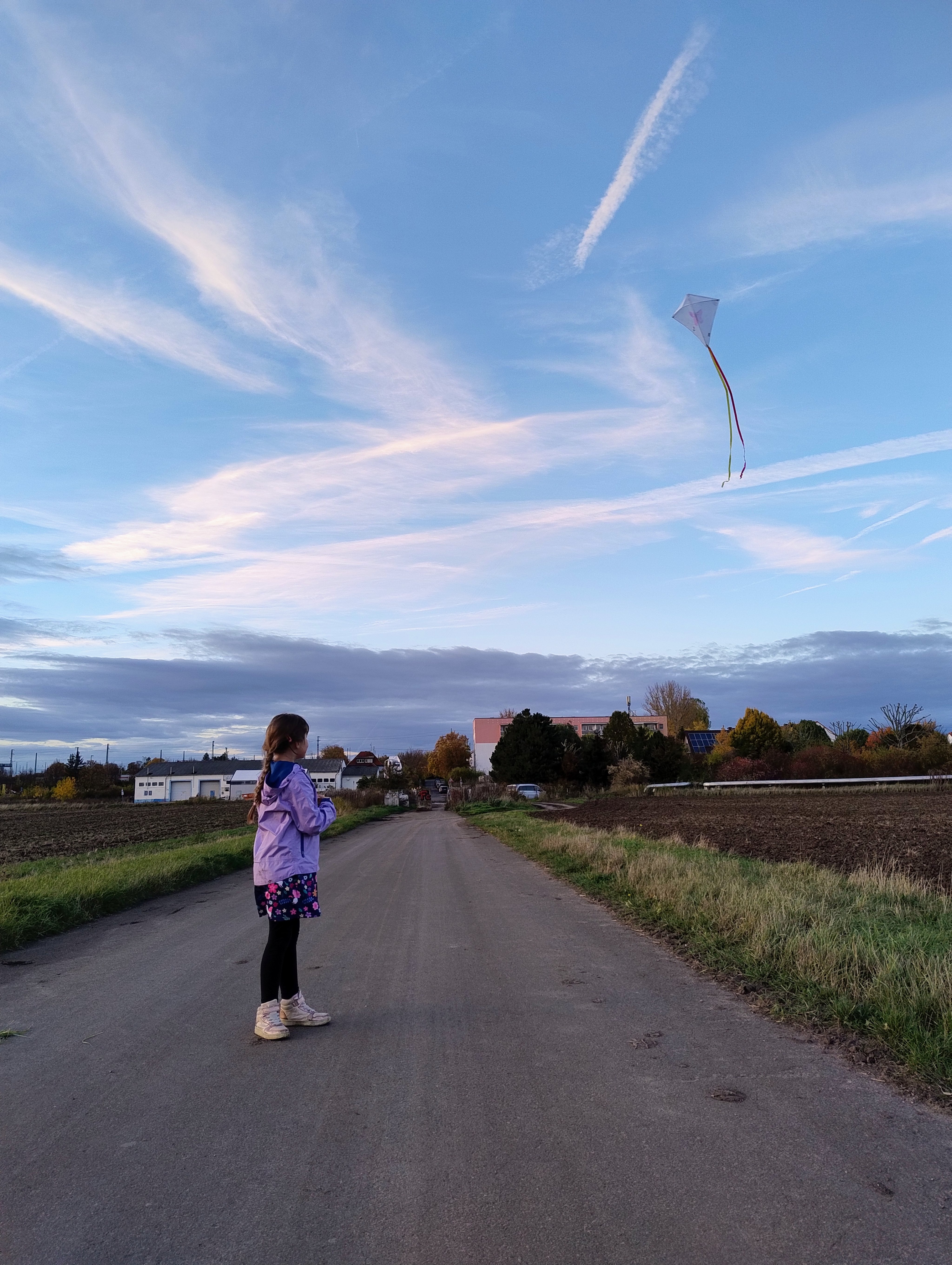Ein Mädchen mit einer lilafarbenen Jacke lässt einen Flugdrachen steigen. Im Hintergrund Häuser