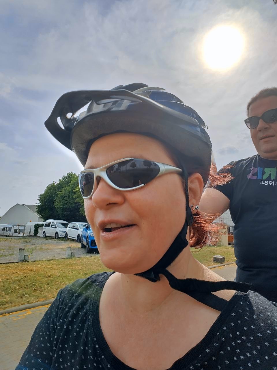 Groß im Vordergrund eine Frau mit Sonnenbrille und Radhelm. Rechts im Hintergrund ein dicker Mann mit Sonnenbrille. Im Hintergrund Straße